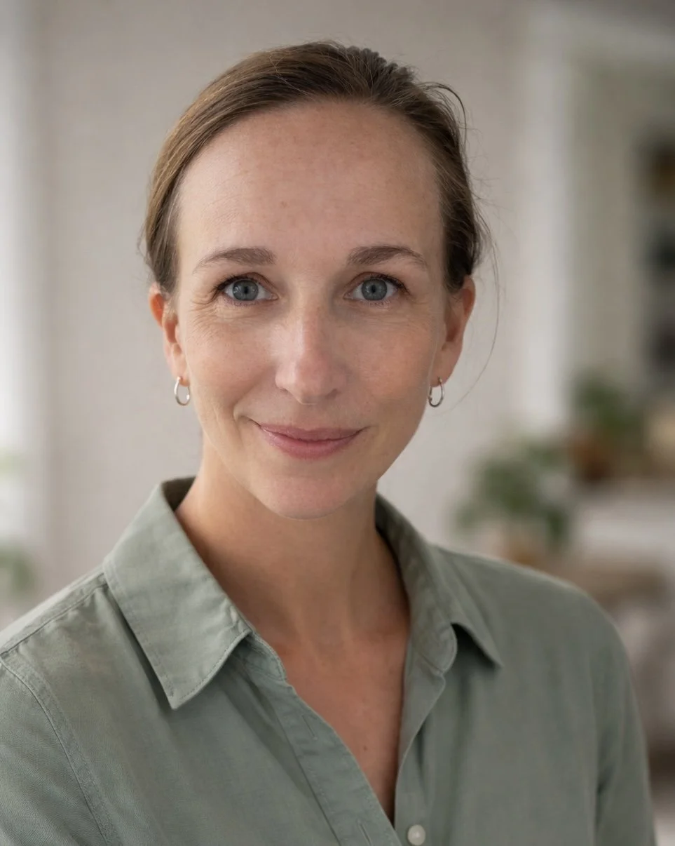 A woman with light skin, blue eyes, and brown hair styled back, wearing hoop earrings and a light green button-up shirt, smiling softly at the camera in a well-lit room.