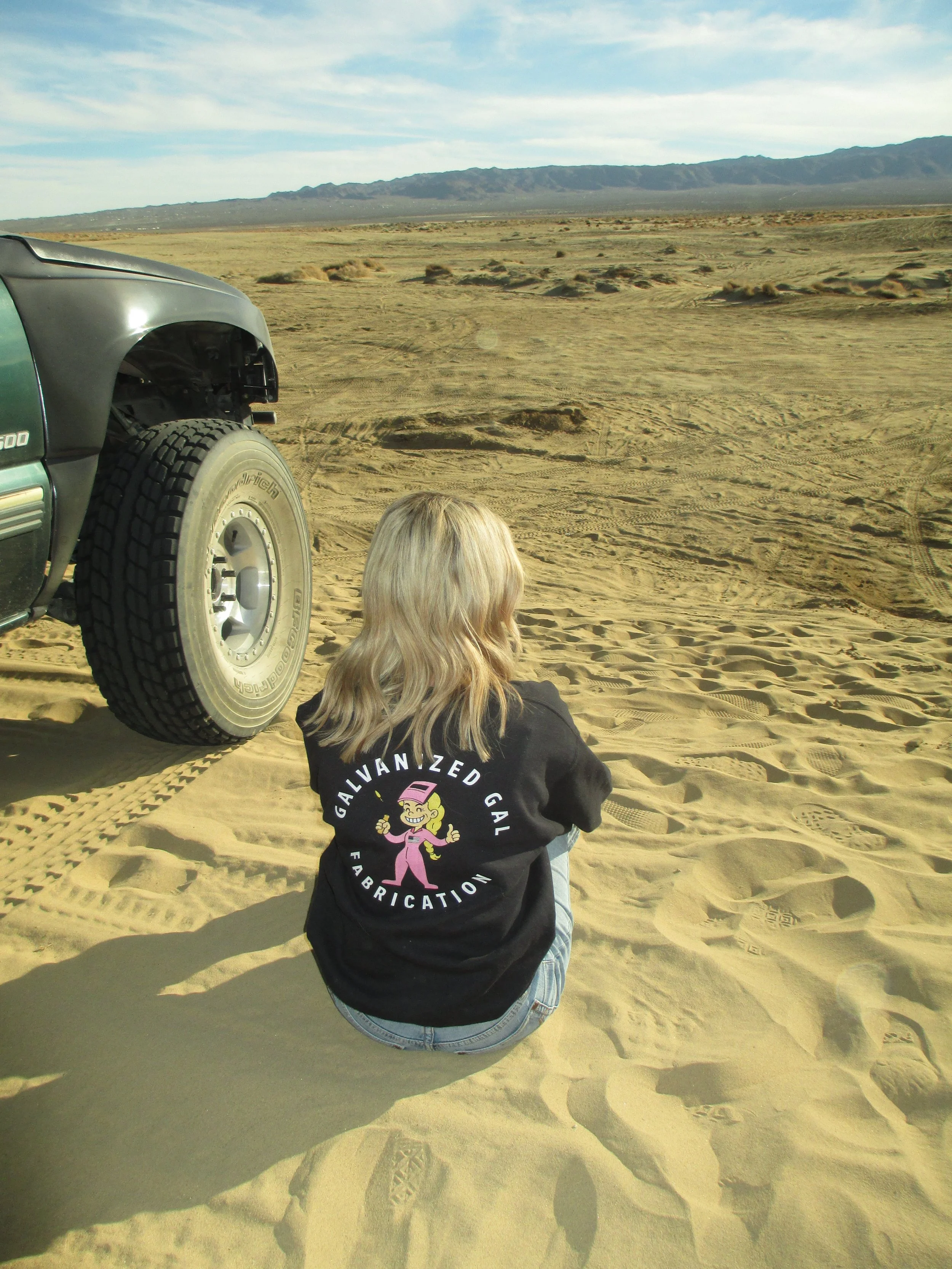 A woman with blonde hair sits on sandy desert ground near a black off-road vehicle, with mountains in the background and a blue sky overhead.