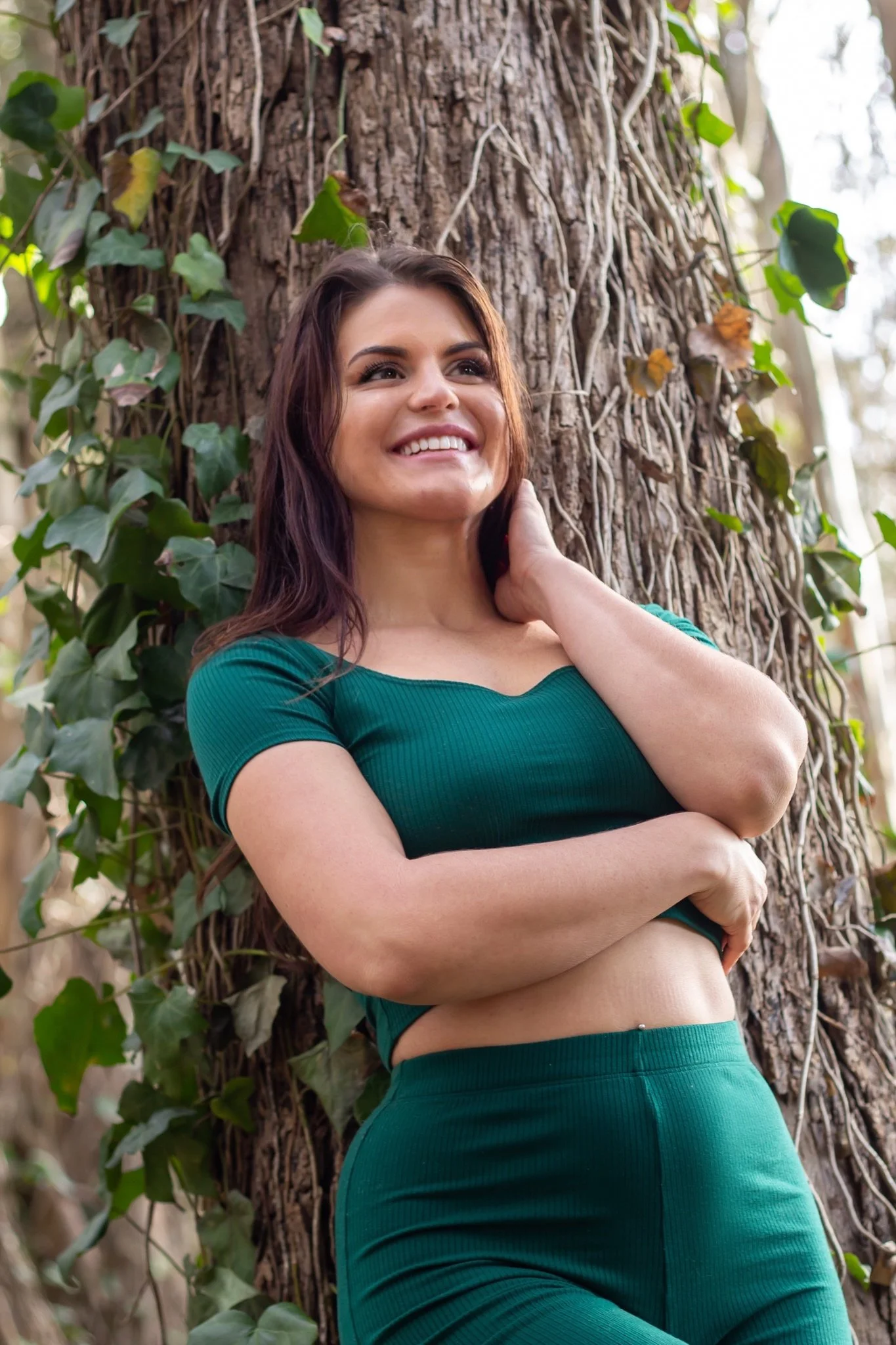 A woman with long brown hair smiling and looking up, leaning against a large tree trunk covered with green ivy in a forested area.