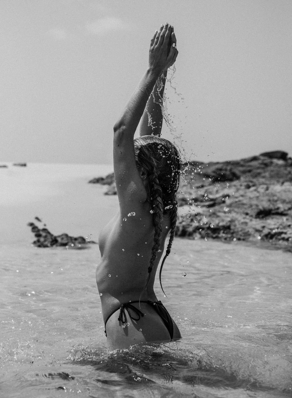 A woman with braided hair standing waist-deep in water, raising her hands above her head, with water splashing around her.