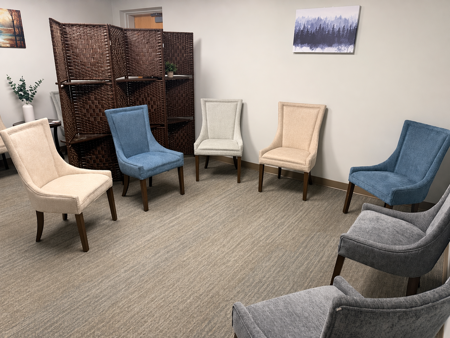 A waiting room with six upholstered chairs in beige, blue, and gray arranged in a semi-circle, with a brown woven room divider behind them and a small table with a plant in a white vase on the left. There are two paintings of nature scenes on the walls.