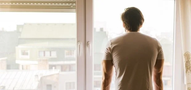 A person standing in front of a window, looking outside at city buildings during daylight.