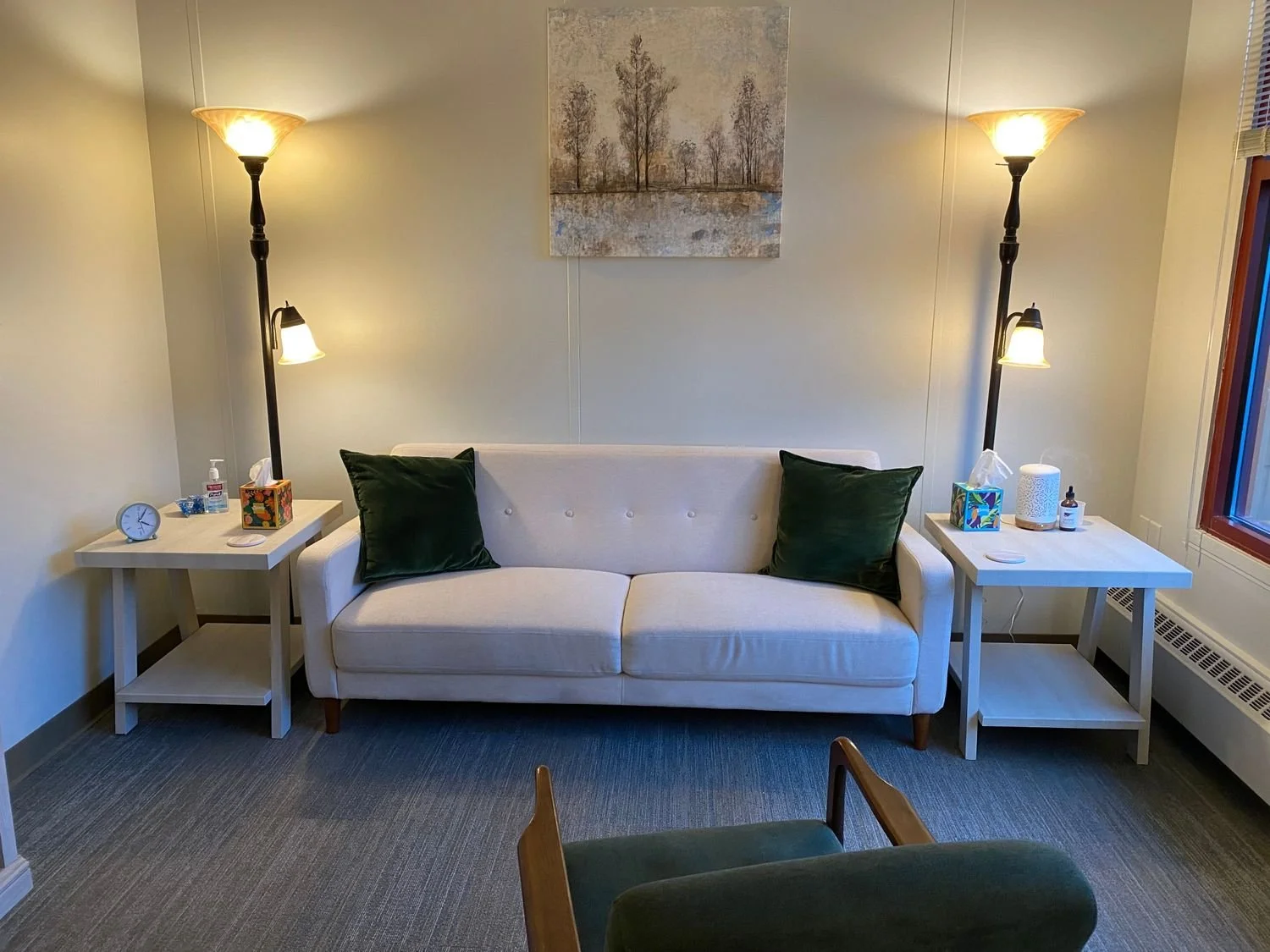 Living room with a white sofa, dark green pillows, two side tables with lamps, artwork of trees on the wall, and a nearby window.