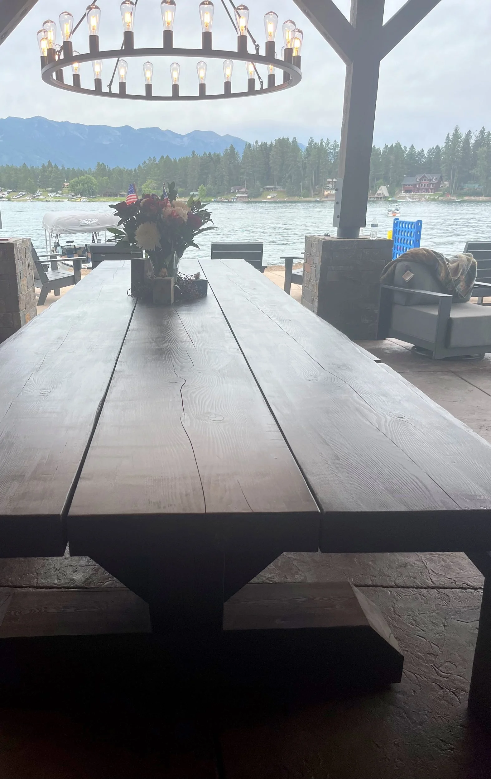 14-foot Douglas fir timber dining table under a timber-frame pavilion, with Echo Lake, a forested shoreline and the Swan Range beyond