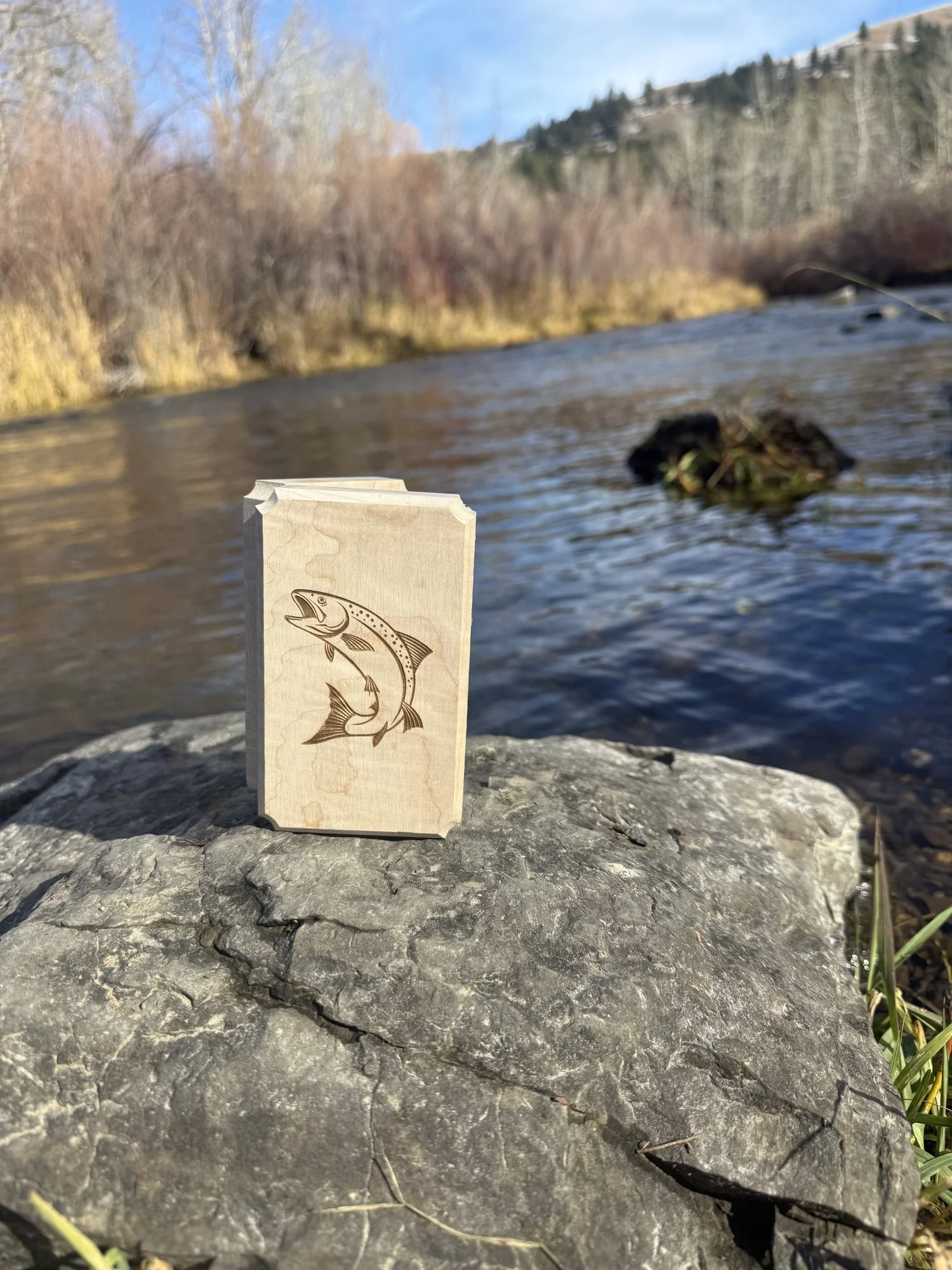 Handmade curly maple fly box resting on river stones beside the Little Blackfoot River in Montana, with hills behind