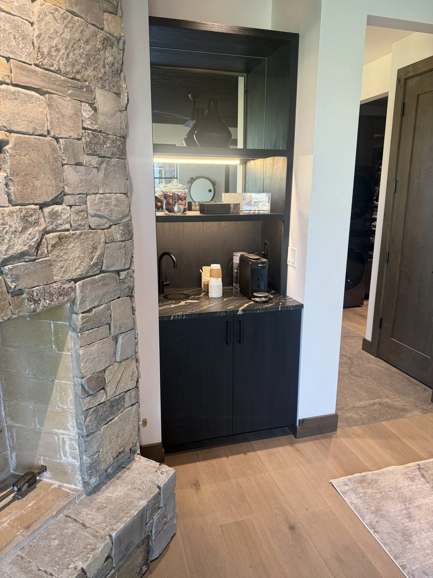 Coffee bar in black-stained red oak with lit open shelving beside a stacked stone fireplace