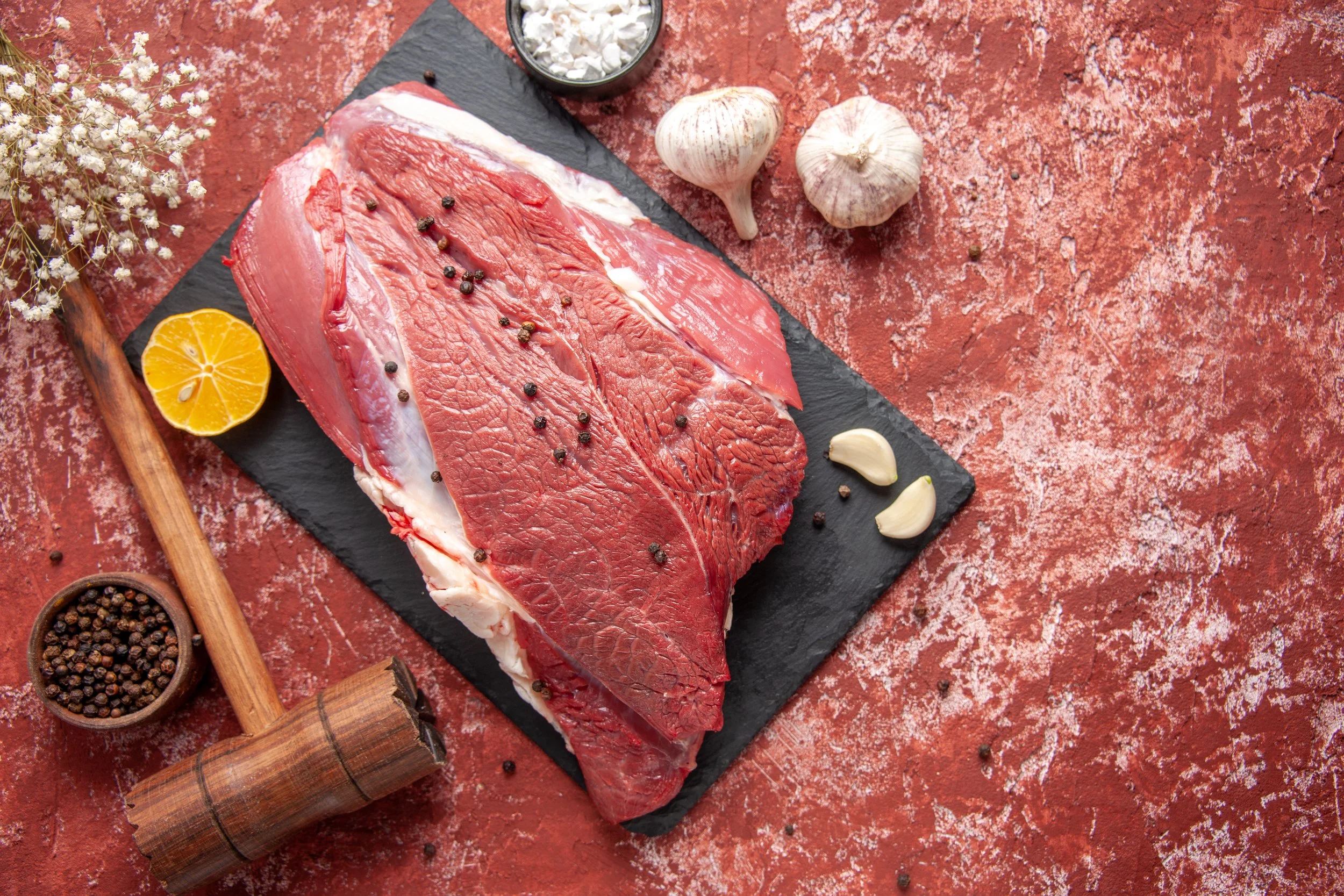 view-fresh-red-meat-with-pepper-black-board-knife-garlics-lemon-spices-brown-wooden-hammer-lemon-oil-pastel-red-background.jpg