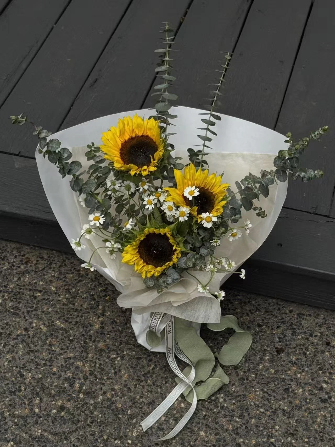 A bouquet of three sunflowers, small white daisies, and Eucalyptus leaves wrapped in white paper and tied with a cream ribbon, resting on a dark wooden surface.
