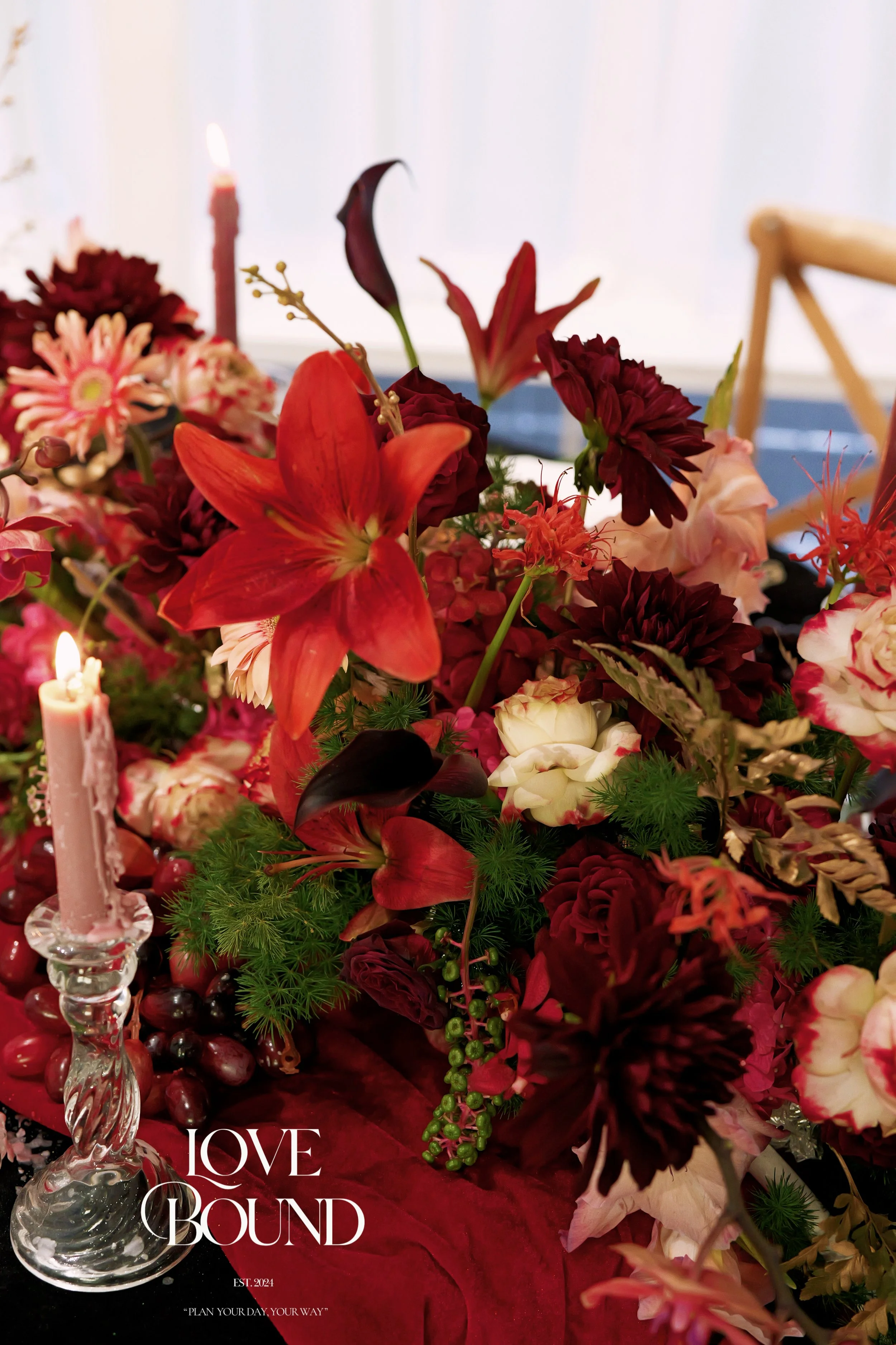 A floral arrangement with red, pink, and white flowers, including lilies, roses, and dahlias, on a table with candles, grapes, and greenery, with the words 'Love Bound' and 'Est. 2024' written on it.