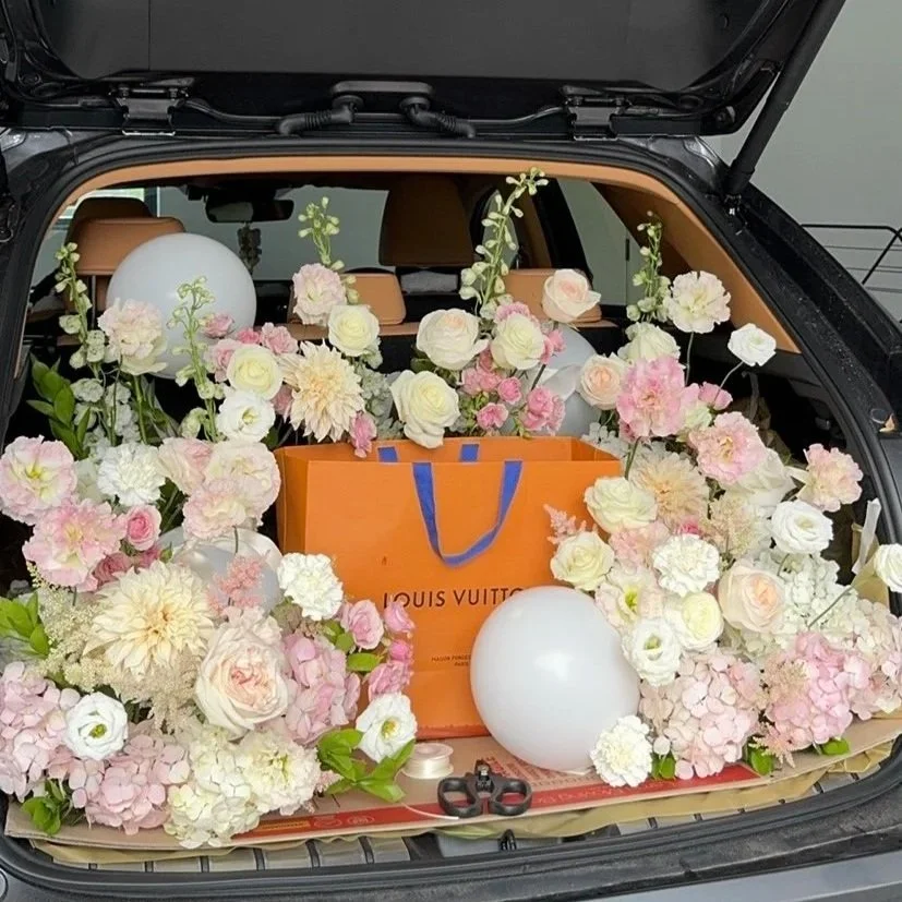 The trunk of a car filled with pink and white flowers, white balloons, an orange Louis Vuitton shopping bag, and scissors.