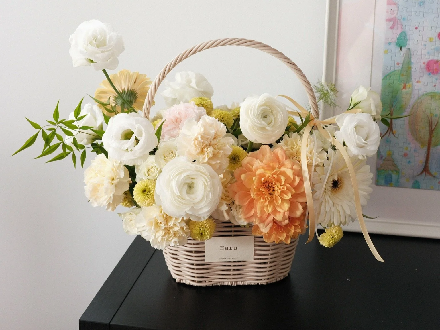 A woven basket with white, peach, and cream flowers, including roses, carnations, and dahlias, with green foliage, on a black table against a white wall.