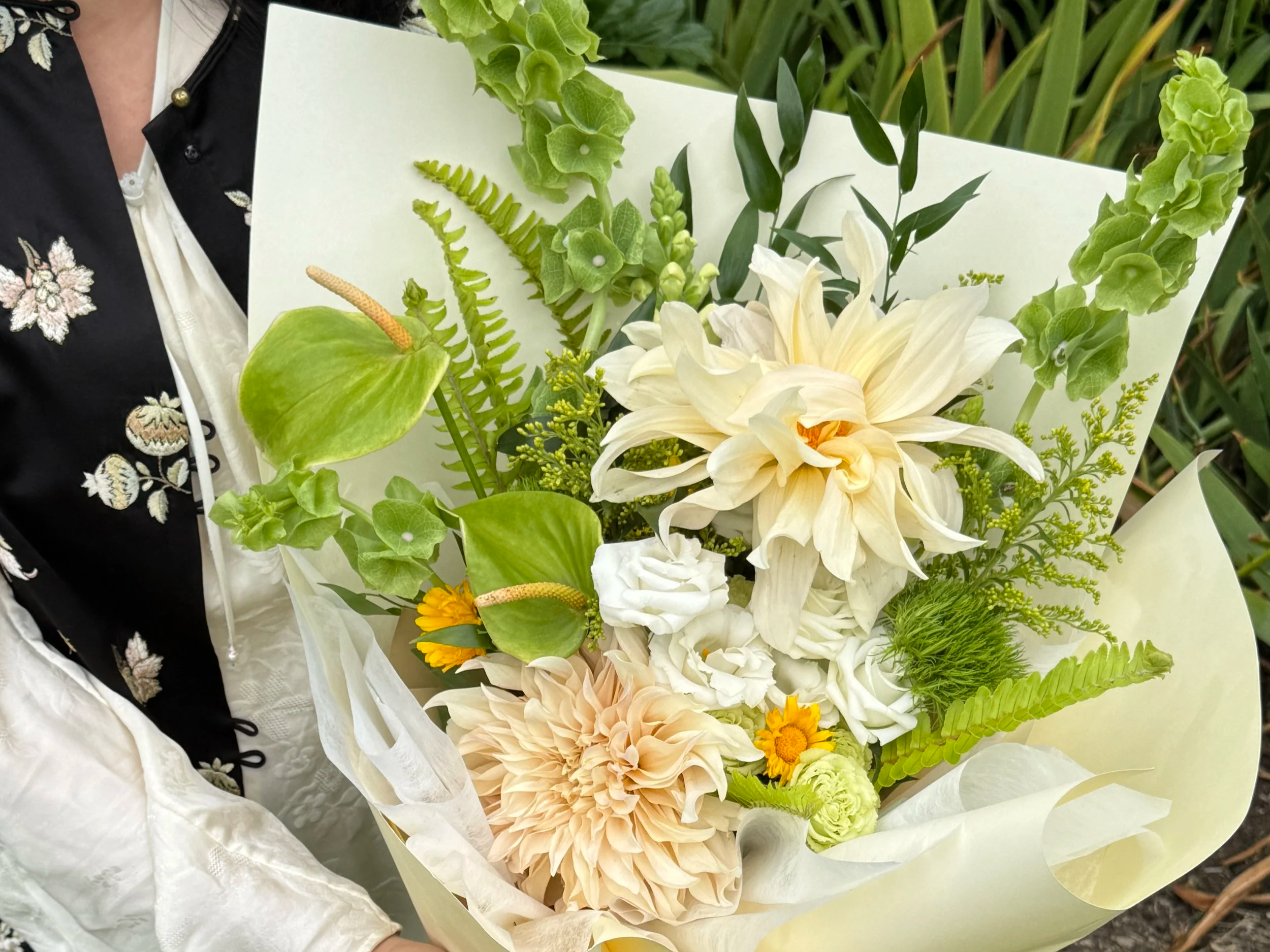 Person holding a large bouquet of white, cream, and yellow flowers, including dahlias, roses, and greenery, wrapped in light-colored paper.