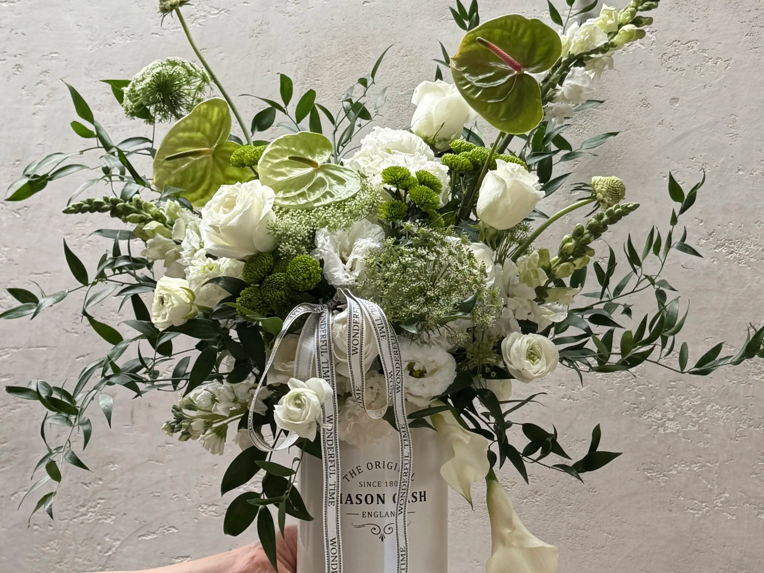 A floral arrangement with white roses, anthuriums, calla lilies, and greenery in a decorative white vase with black text, accented with a black and white ribbon.