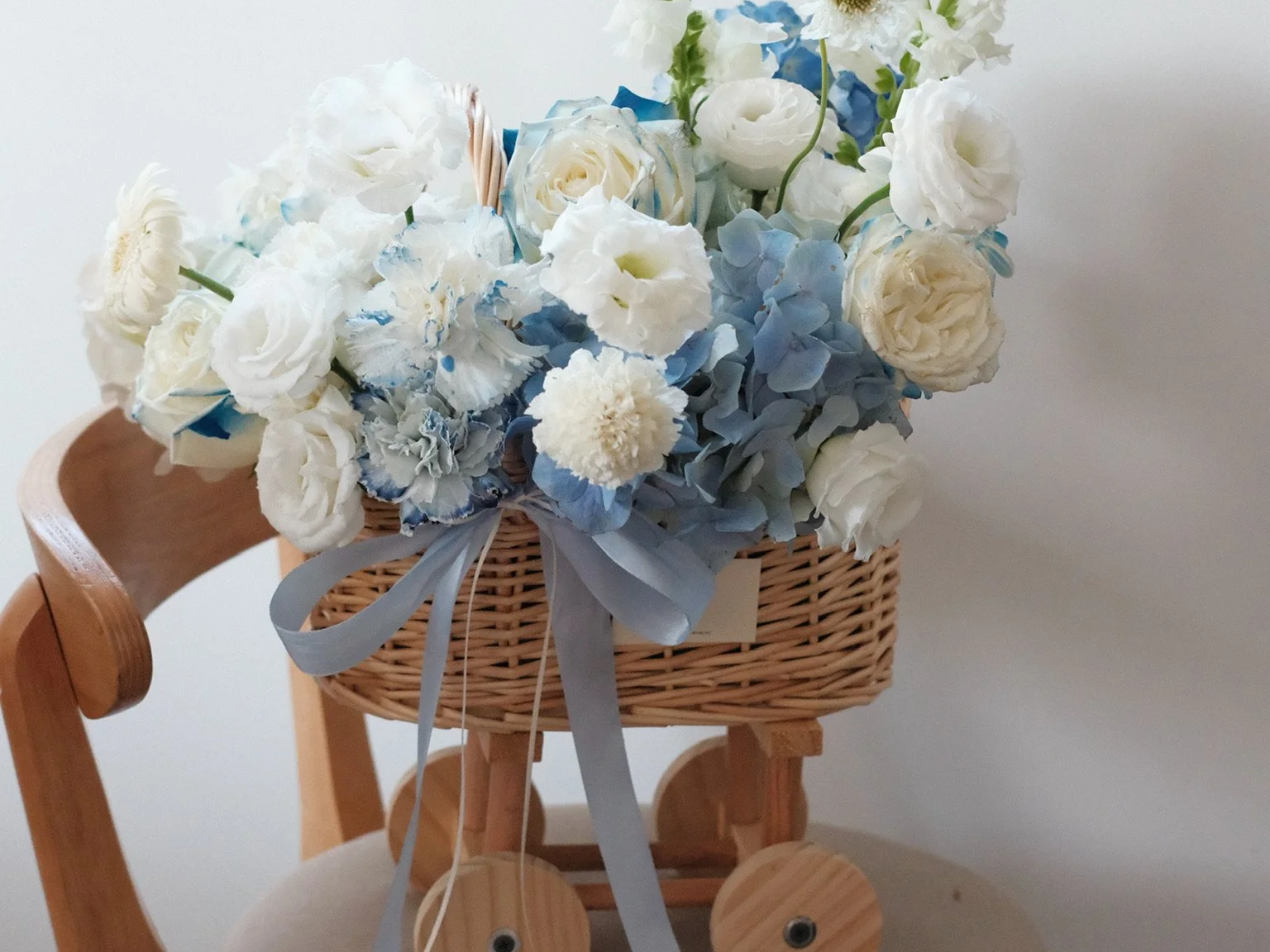A wicker basket filled with white and blue flowers, including roses and hydrangeas, placed on a wooden chair with a ribbon bow.