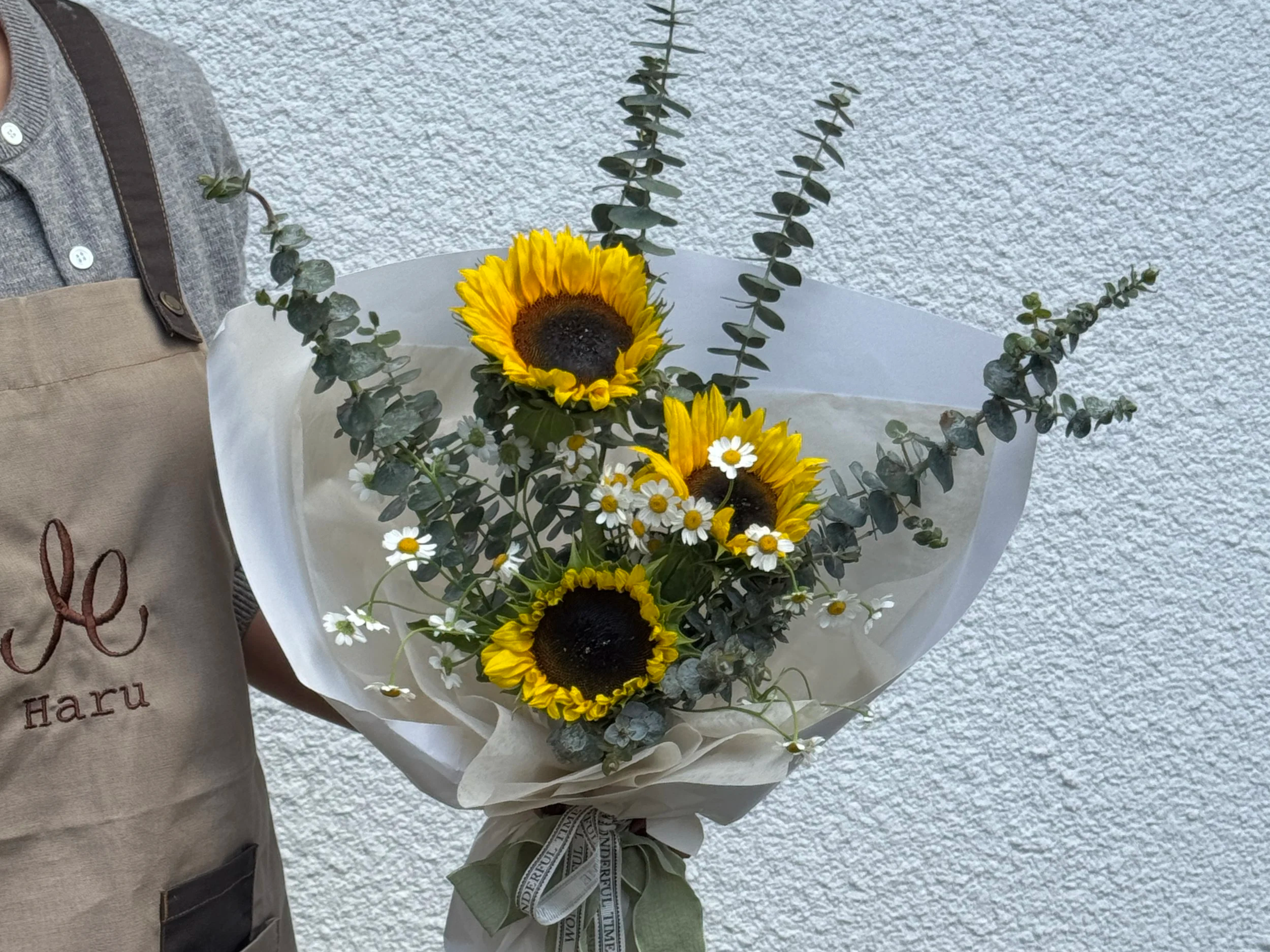 Bouquet of sunflowers, small white daisies, and eucalyptus leaves wrapped in white paper, held by a person wearing a beige apron and gray shirt.