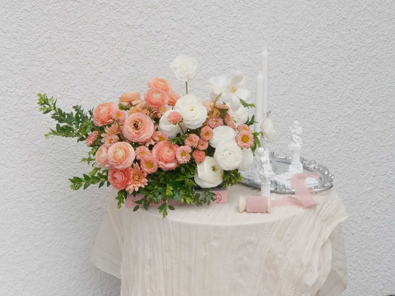 Floral arrangement of pink and white flowers on a draped table next to white candles and decorative silver tray.