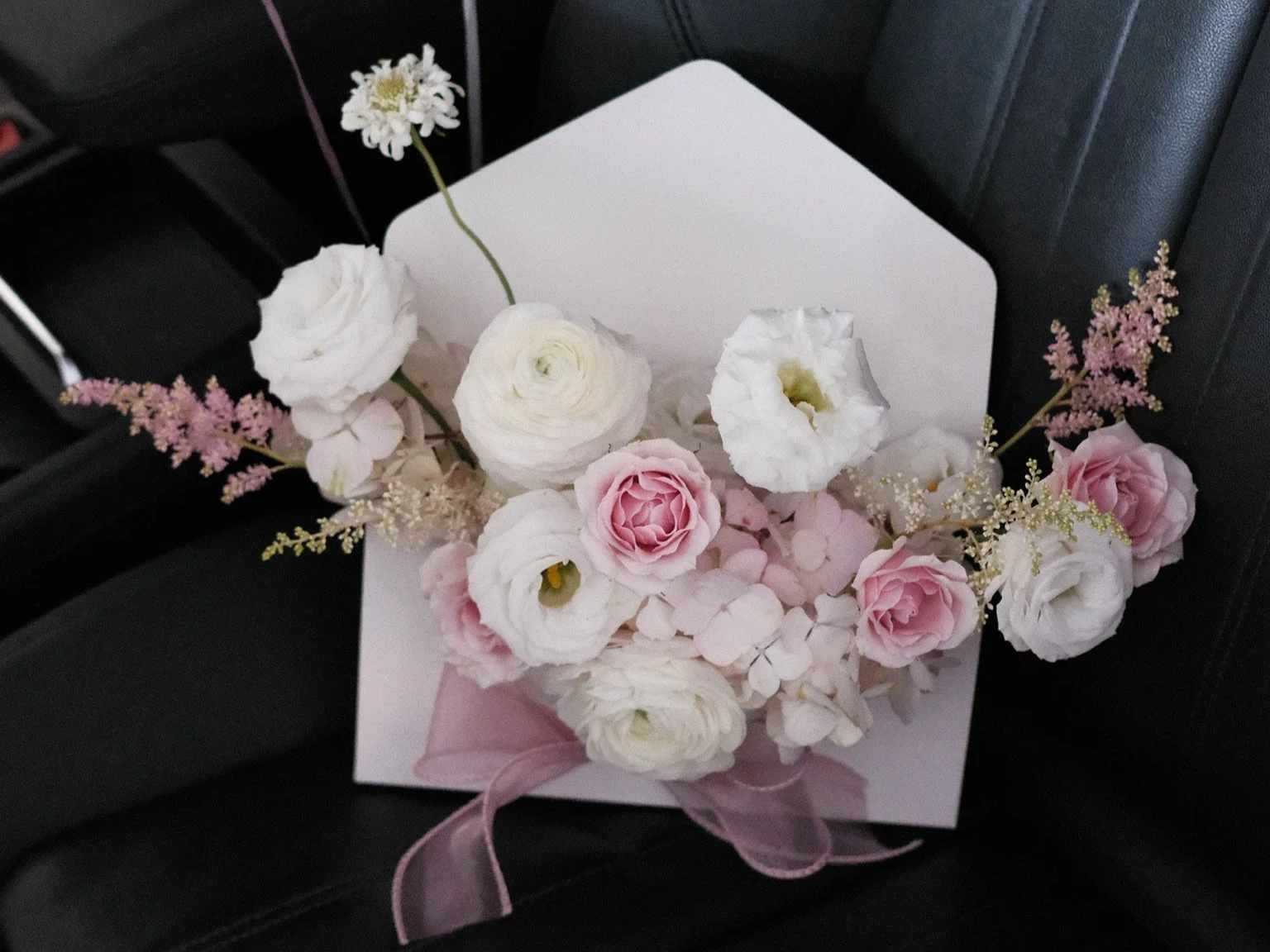 A bouquet of pink and white flowers, including roses and lisianthus, arranged with pink hydrangeas and decorative greenery, wrapped with a pink ribbon, placed on a black leather chair.