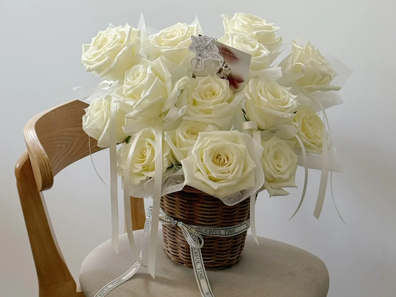 A basket full of white roses with white ribbon and a gift card, placed on a cushioned chair.