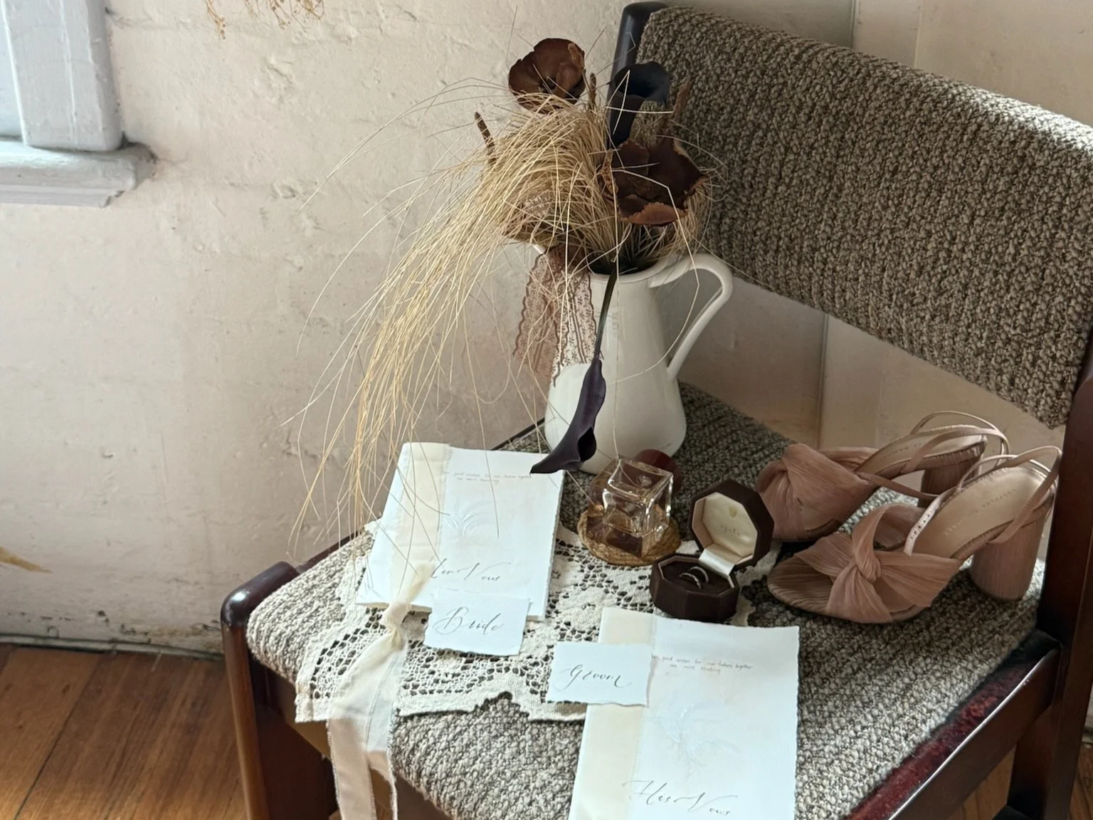Wedding accessories on a chair, including a white lace cloth, a pair of pink high heels with bows, a ring box with a ring, dried flowers in a white pitcher, and handwritten notes labeled 'Bride' and 'Groom'.