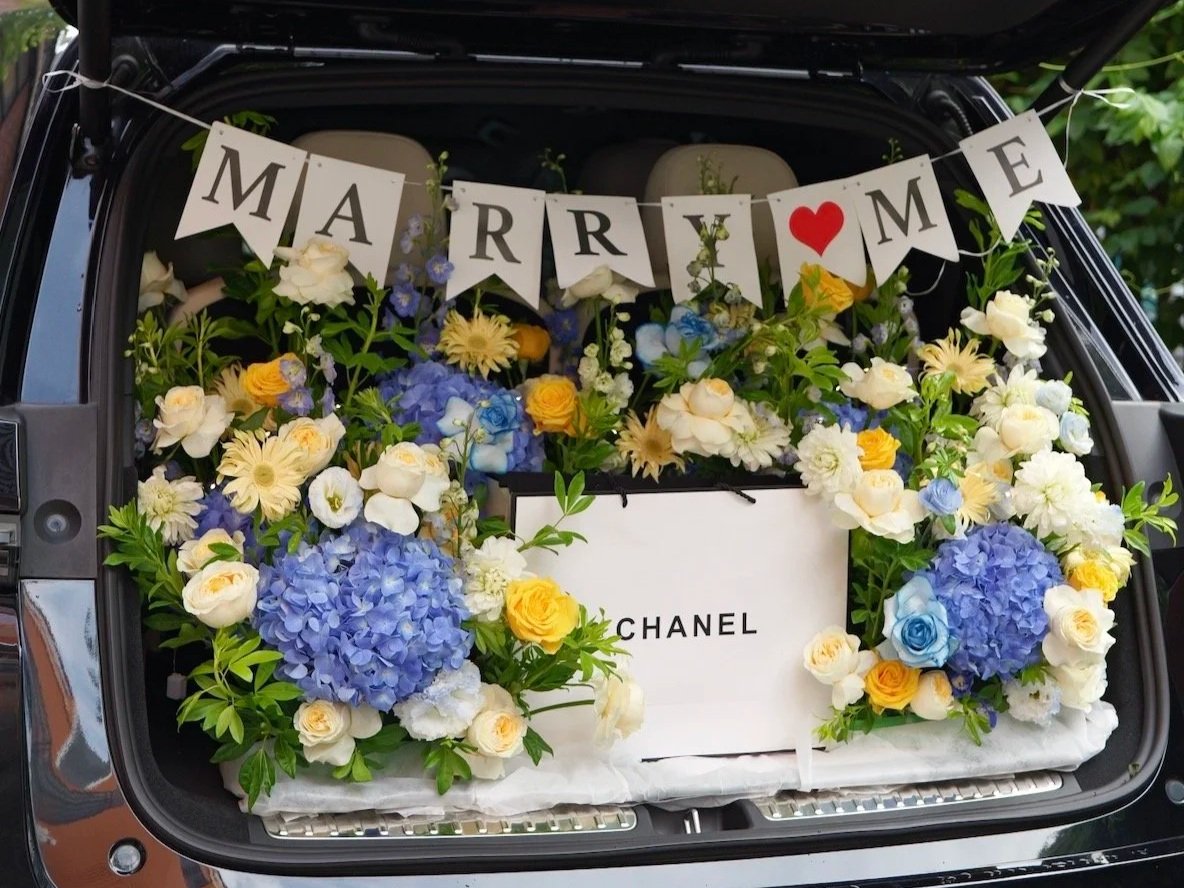 The open trunk of a car filled with a large arrangement of white, yellow, purple, and blue flowers, a Chanel shopping bag, and a banner that spells out "MARRIAGE" with a heart symbol replacing the "I".
