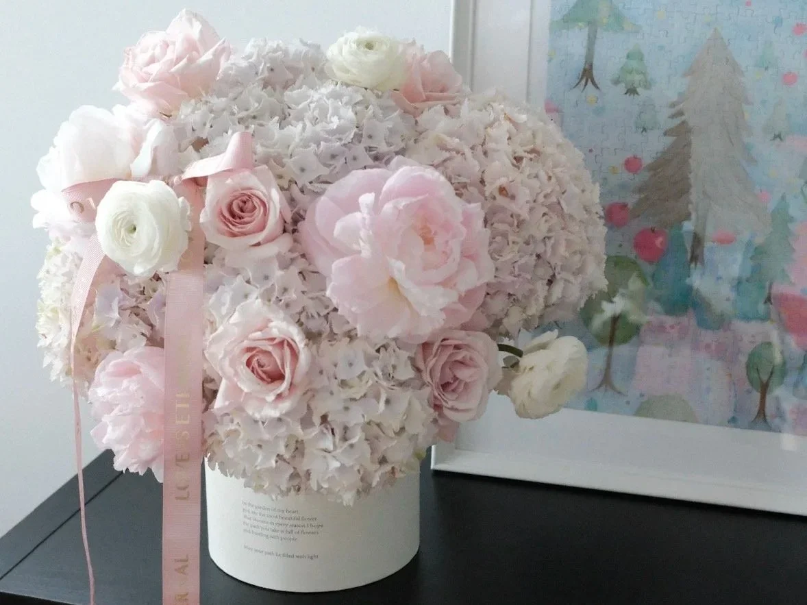 A bouquet of light pink and white flowers, including roses and peonies, arranged in a white container with a pink ribbon attached, placed on a black surface next to a framed colorful illustration of trees.