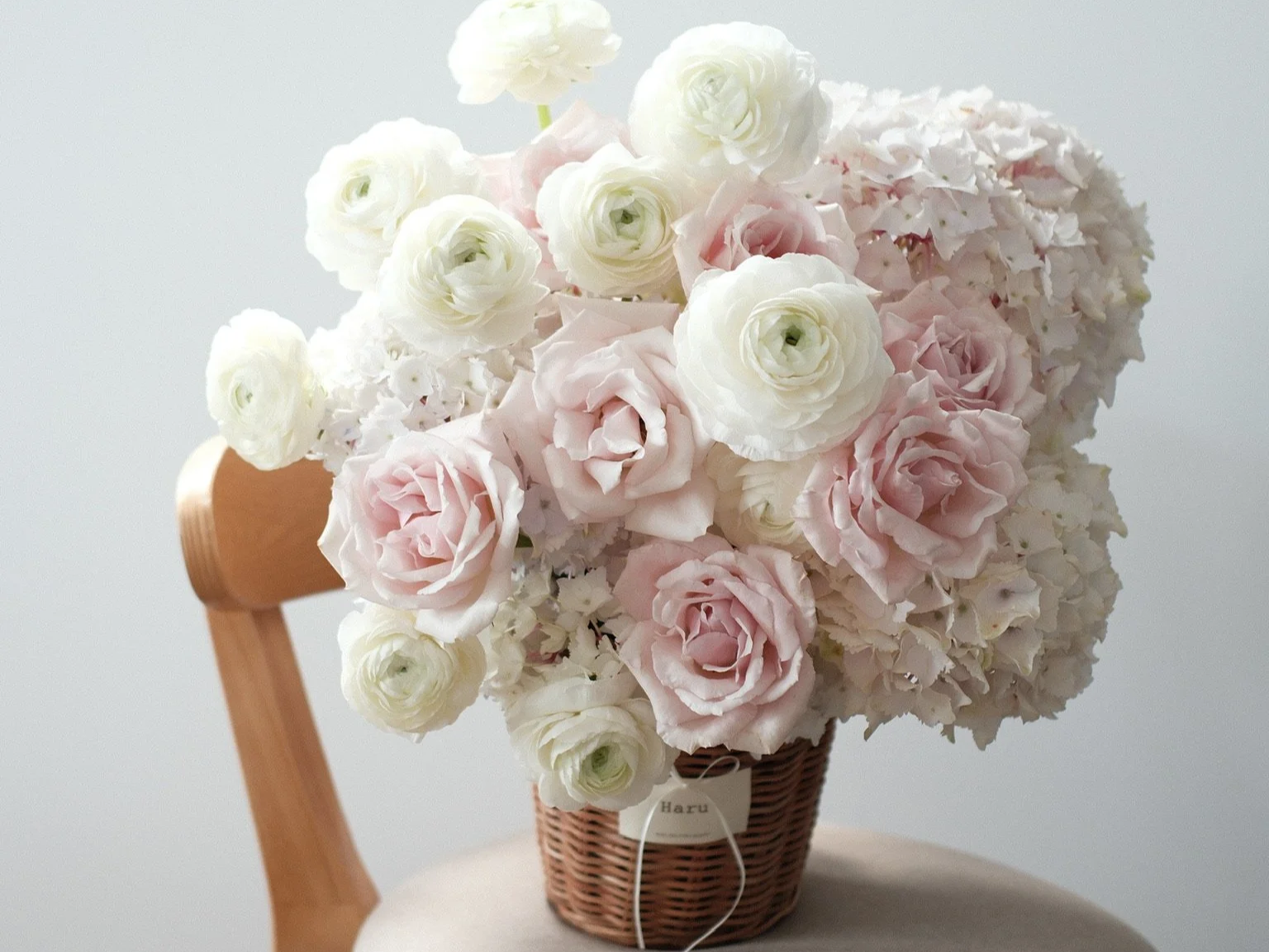 A wicker basket filled with white and light pink roses, sitting on a rounded surface next to a wooden chair back, against a light grey background.