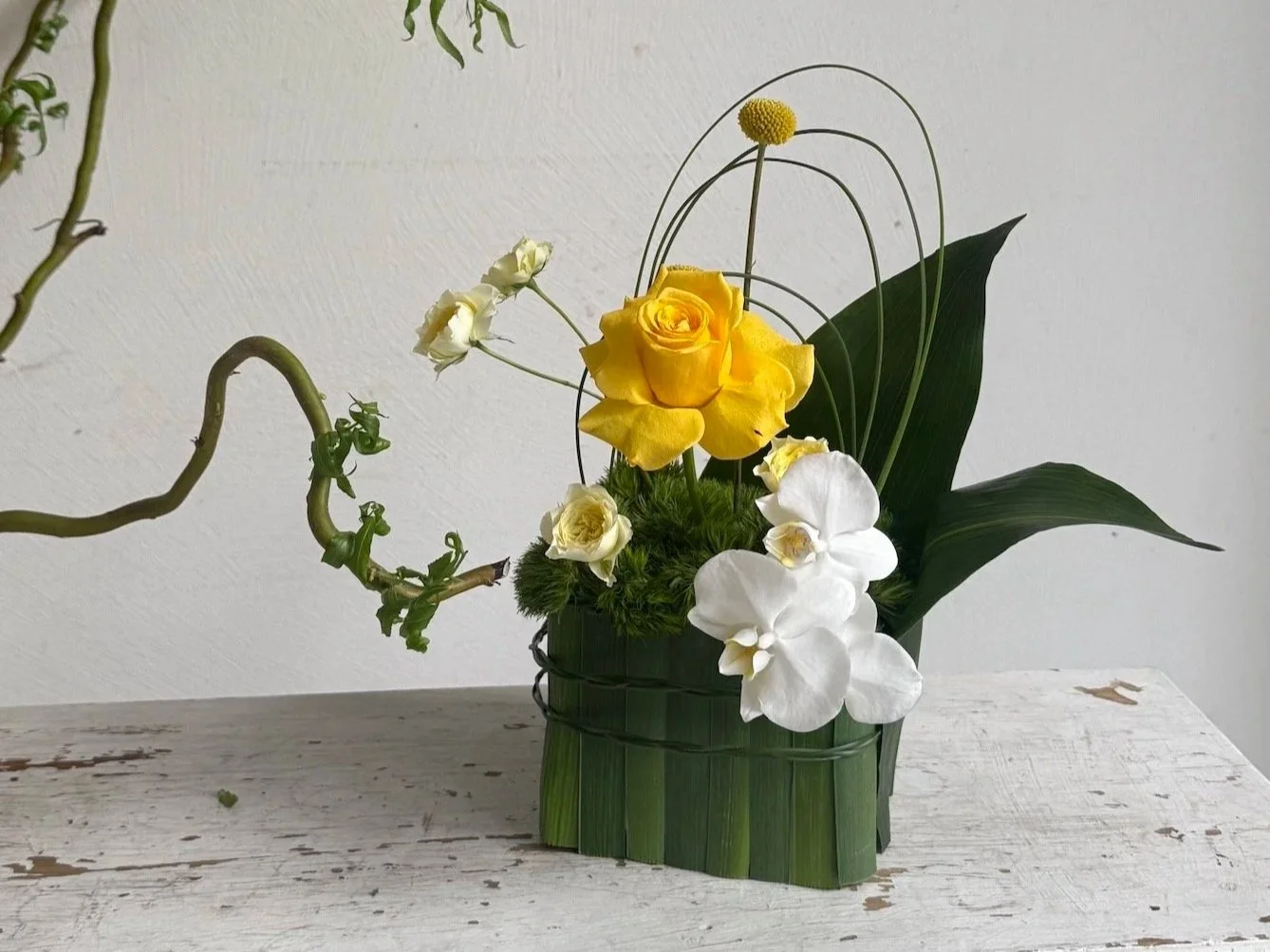 Arrangement of yellow roses, white orchids, and green foliage in a green woven basket.