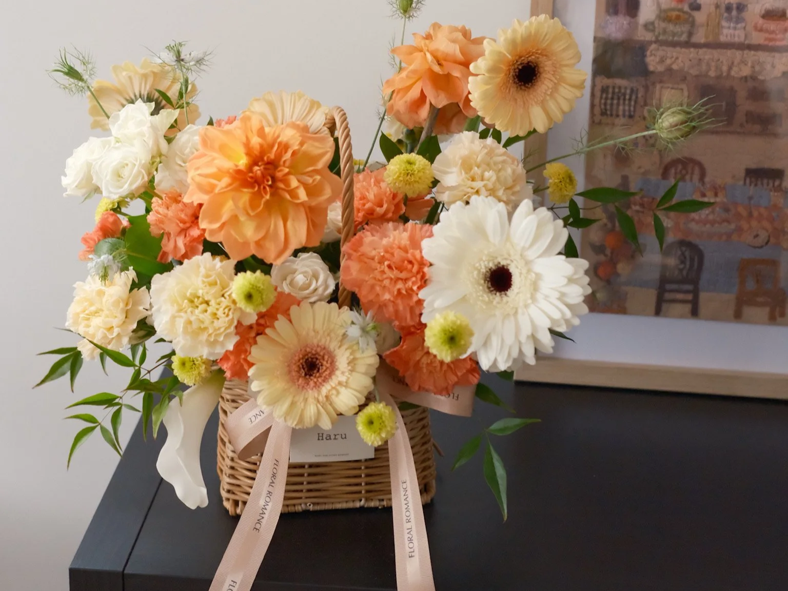 A wicker basket filled with peach, cream, and white flowers, decorated with a pink ribbon that has the words "FLORAL ROMANCE" printed on it.
