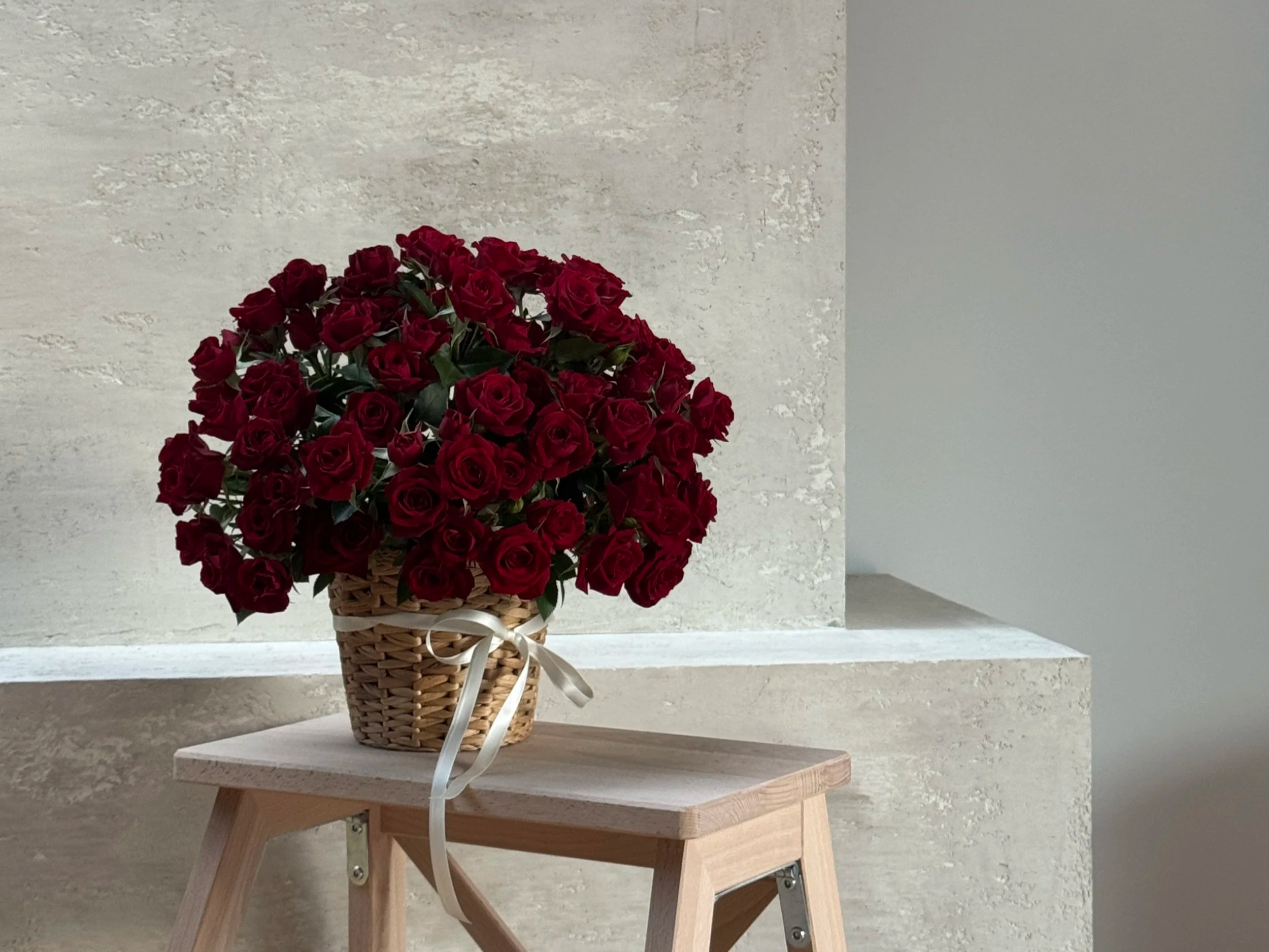 A basket filled with red roses on a small wooden table against a textured wall.
