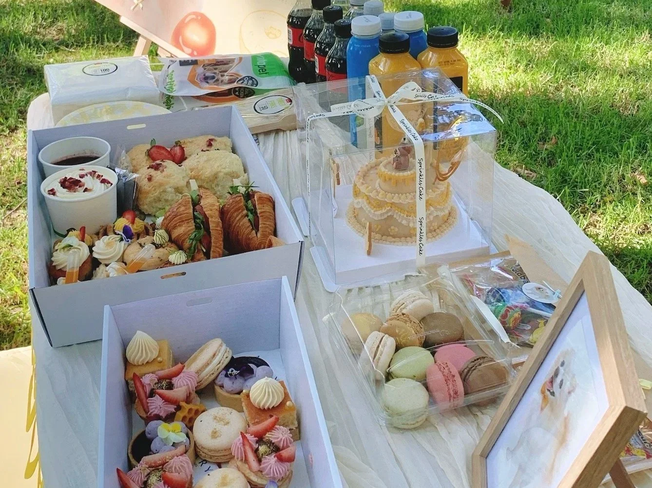 A table set outdoors with various desserts including macarons, cupcakes, and a cake, along with boxes of pastries and bottles of beverages.