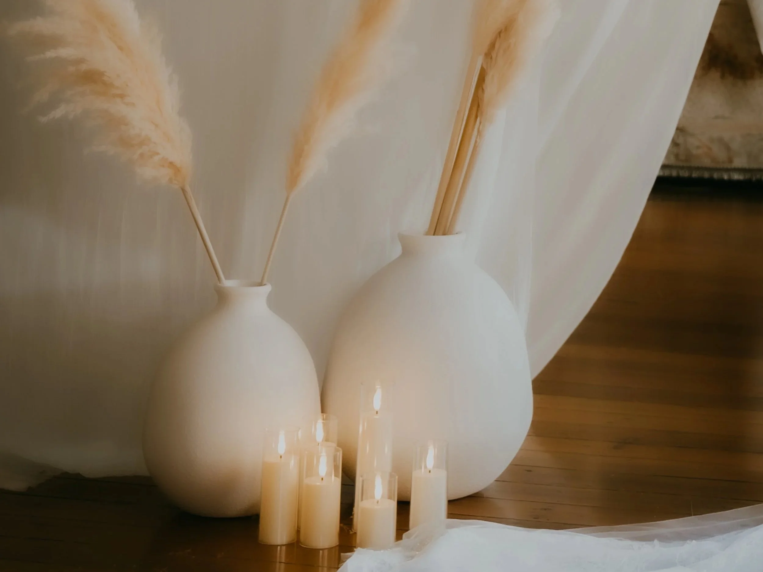 Two white ceramic vases with dried pampas grass and reed sticks, surrounded by small white flameless candles on a wooden floor, with a light-colored draped fabric in the background.