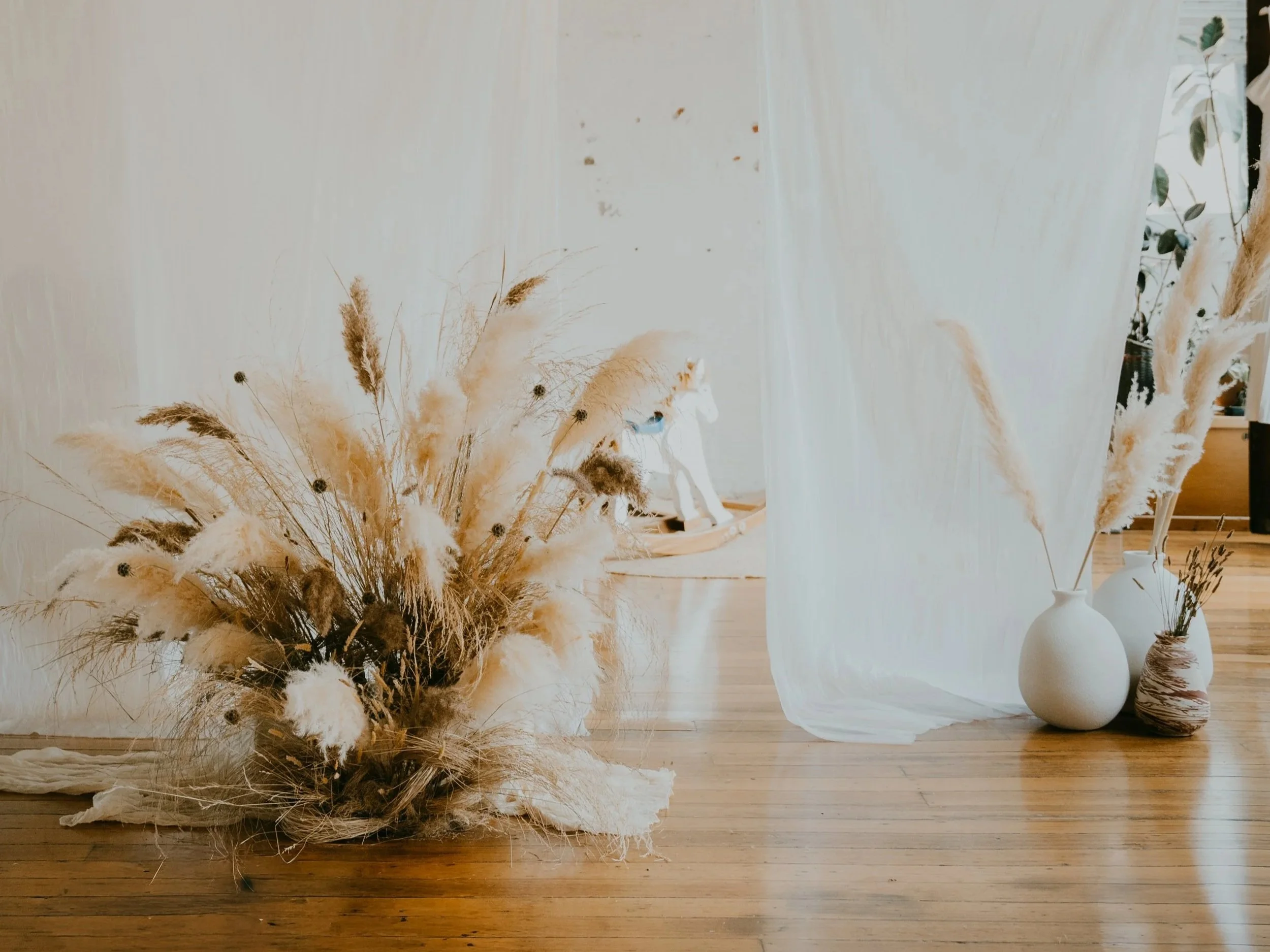 Arrangement of dried pampas grass and other dried flowers in vases on a wooden floor, next to a sheer white curtain.