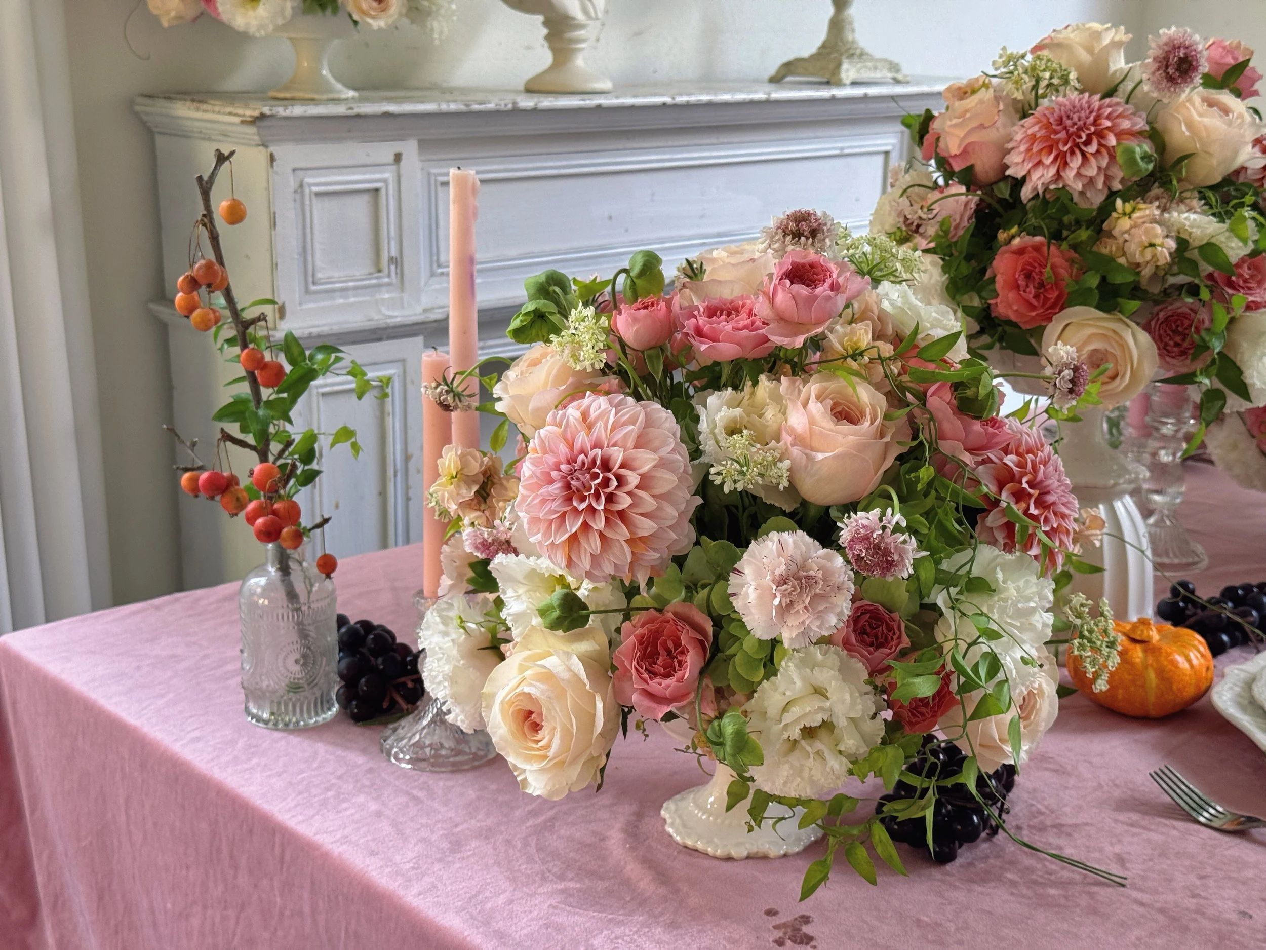 A pink tablecloth with floral arrangements, candles, and small decorative pumpkins on a decorated table in a room with ornate white furniture and a light-colored wall.