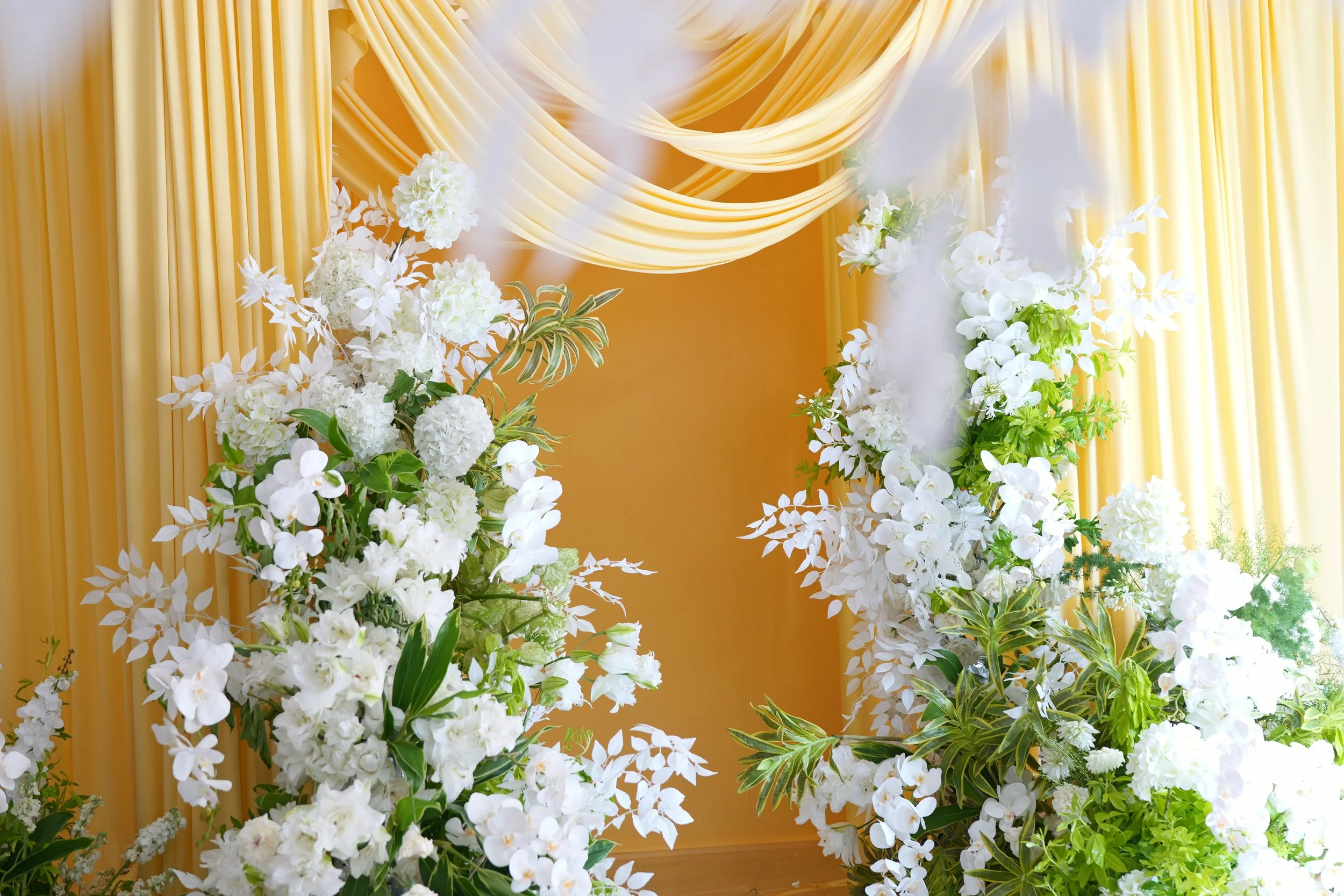 White floral arrangement with greenery set against yellow drapes and a yellow wall, suitable for a wedding or special event.
