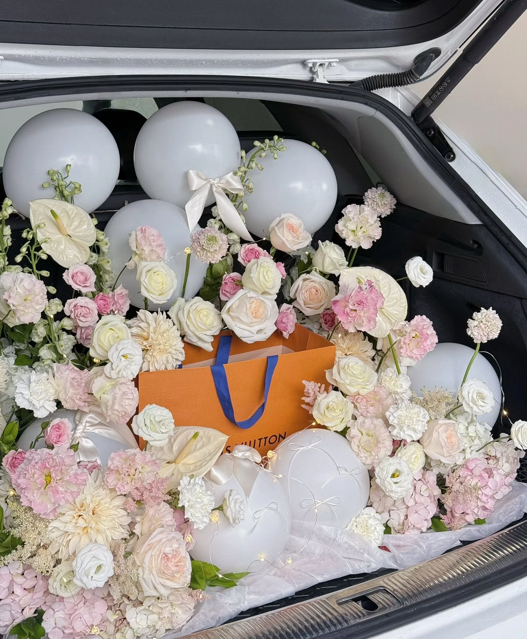 A car trunk filled with pink and white flowers, white balloons, a Louis Vuitton shopping bag, and decorative string lights.