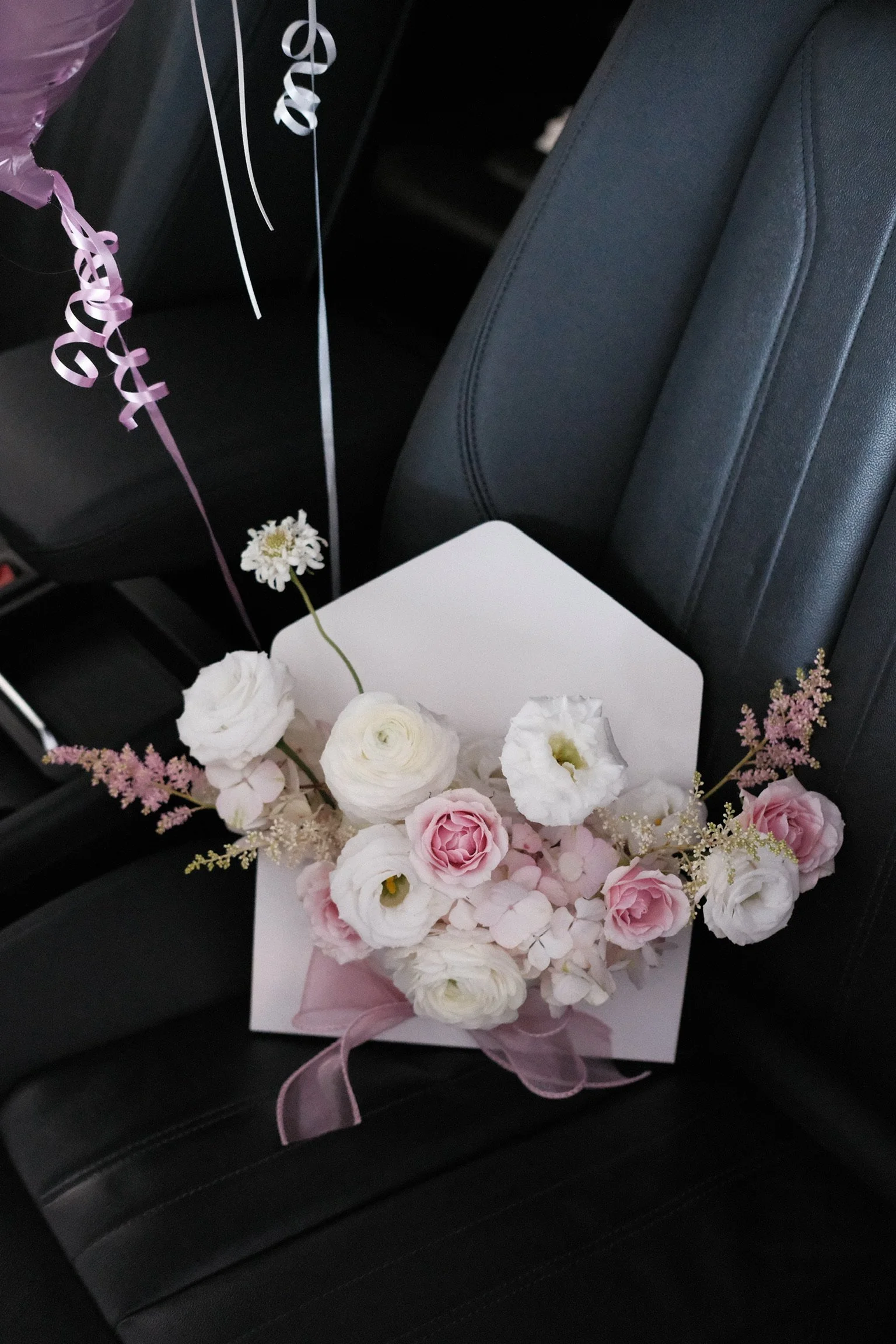 A white box containing a bouquet of pink and white flowers with pink ribbons and curly ribbons, sitting on a black car seat.