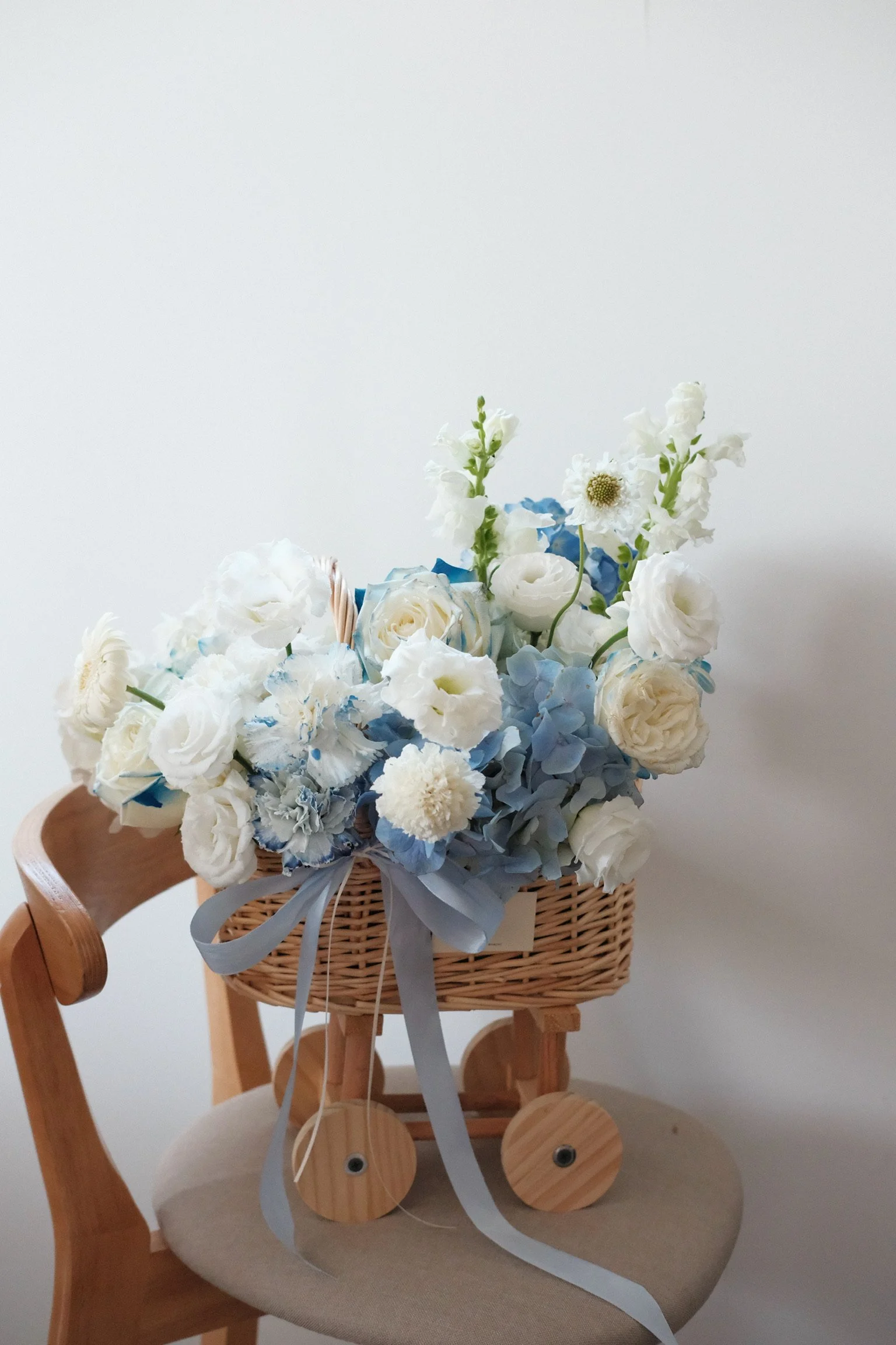 A wicker flower basket filled with white and light blue flowers placed on a wooden chair with a light-colored seat, against a plain white wall.