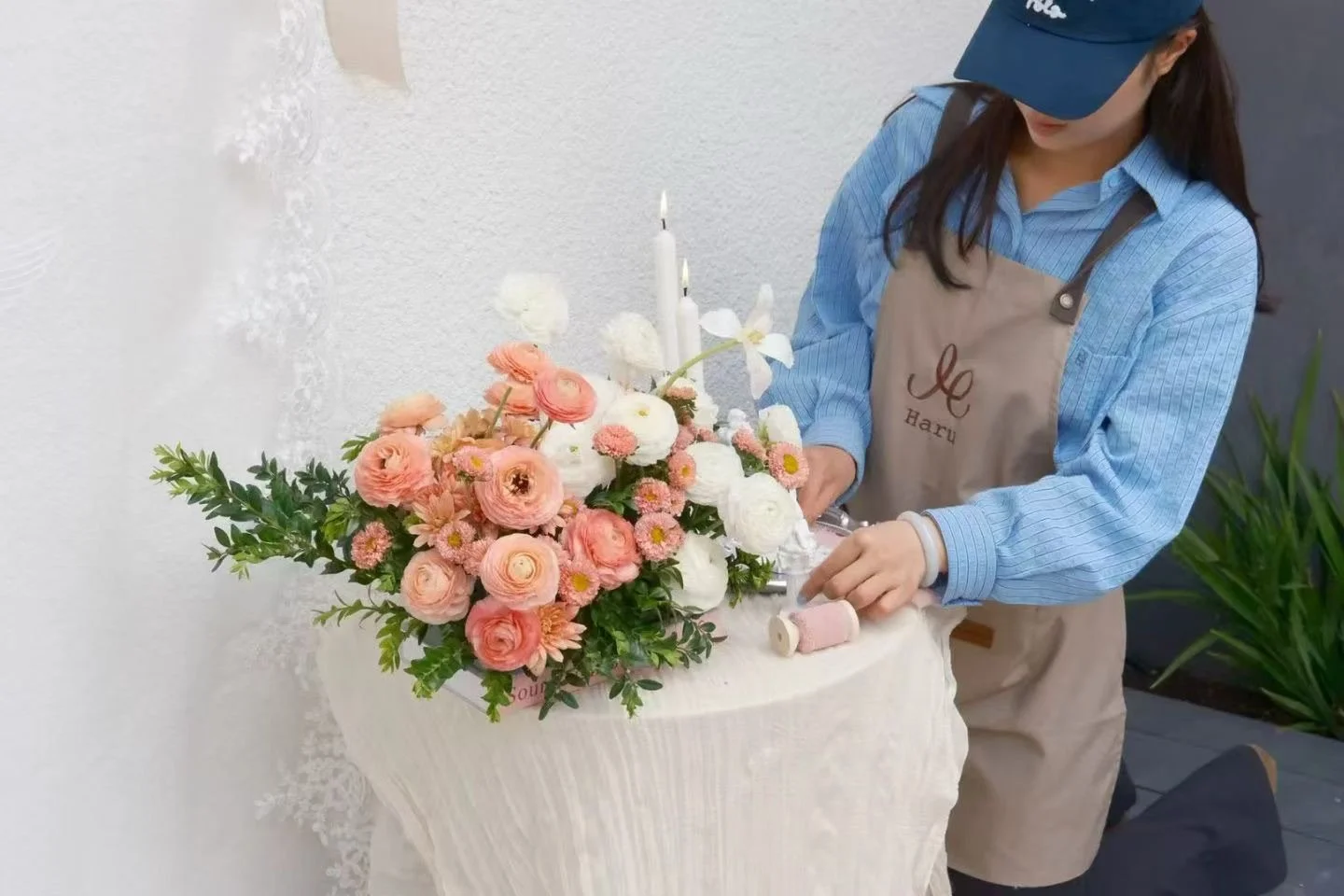 A woman arranging a floral display with pink and white flowers, candles, and greenery on a beige table, wearing a blue shirt, a beige apron with a monogram, and a blue cap.