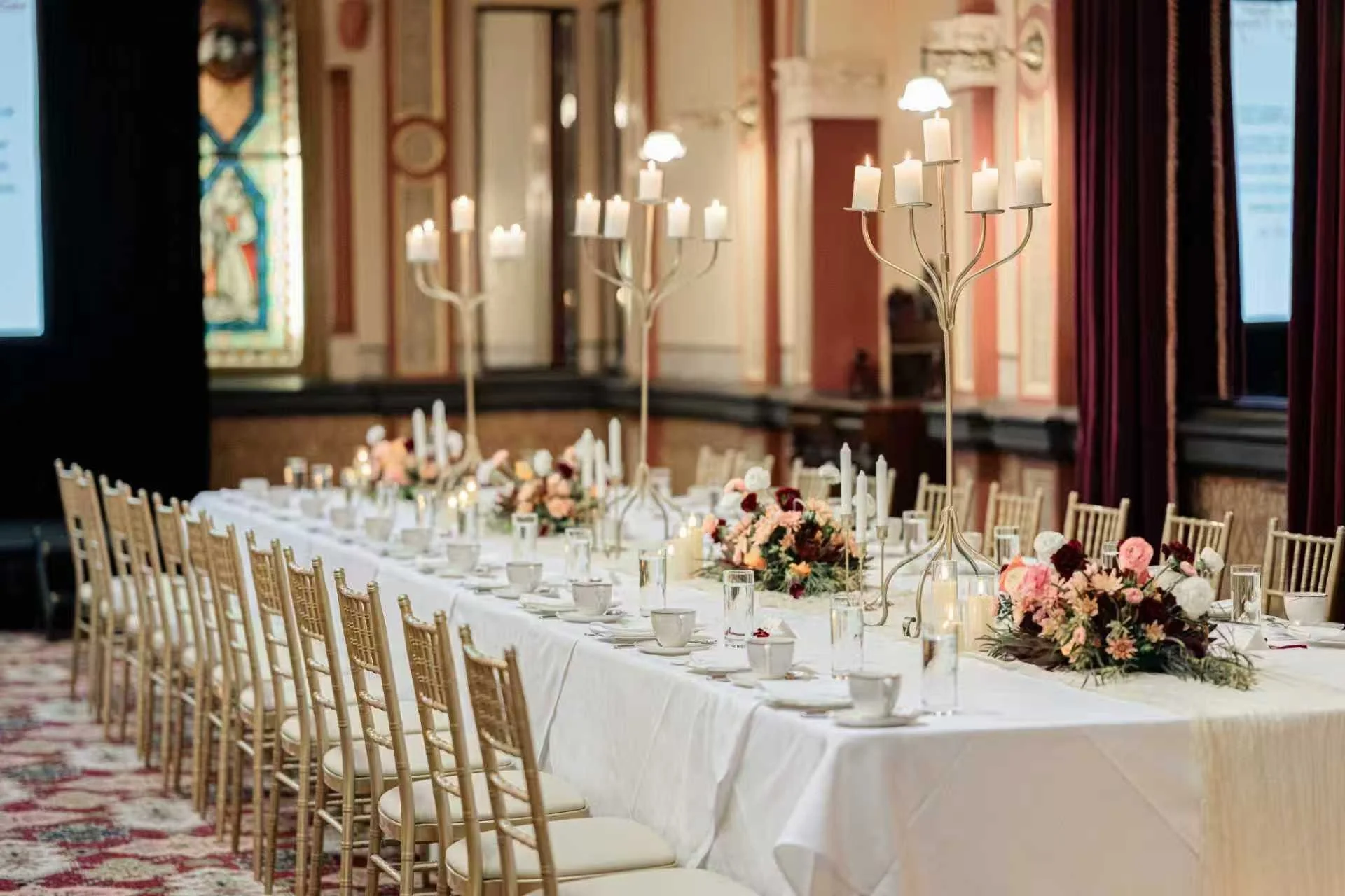 Elegant banquet table set up with floral centerpieces, candles, and candelabras in a decorated room with stained glass window and red curtains.