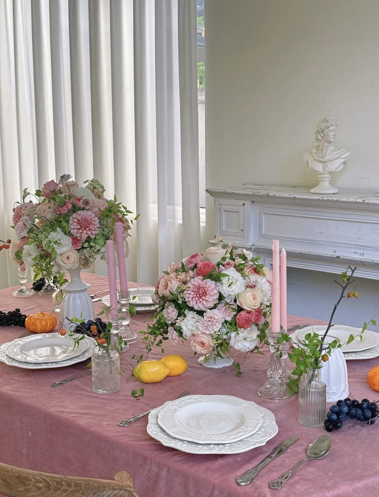 Elegant dining table set with pink floral arrangements, pink candles, fruit, and fine china, in a bright room with white curtains and classical bust sculpture on a white mantel.