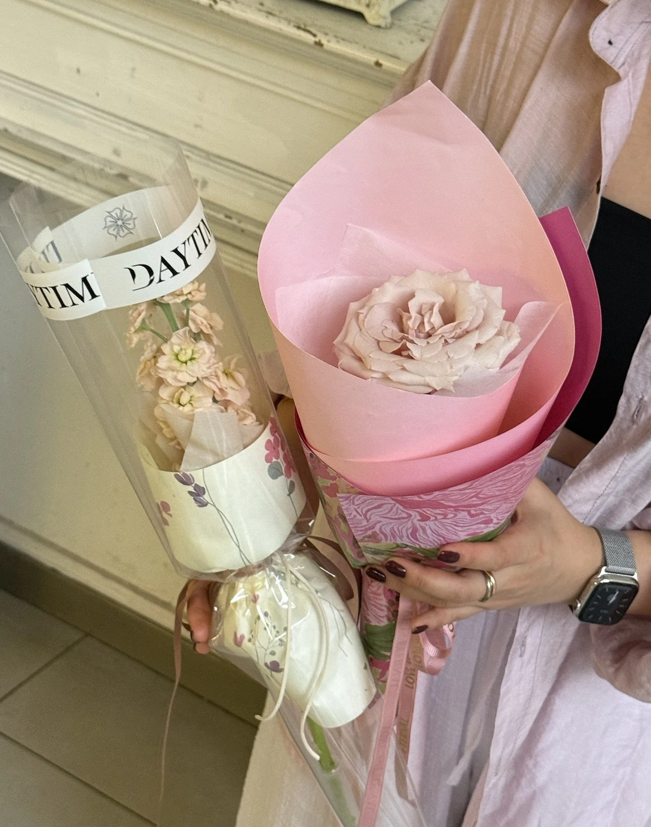 A person holding two wrapped flower bouquets, one in pink wrapping with a large pale flower, and the other in clear wrapping with pink flowers, both wrapped with pink ribbons.