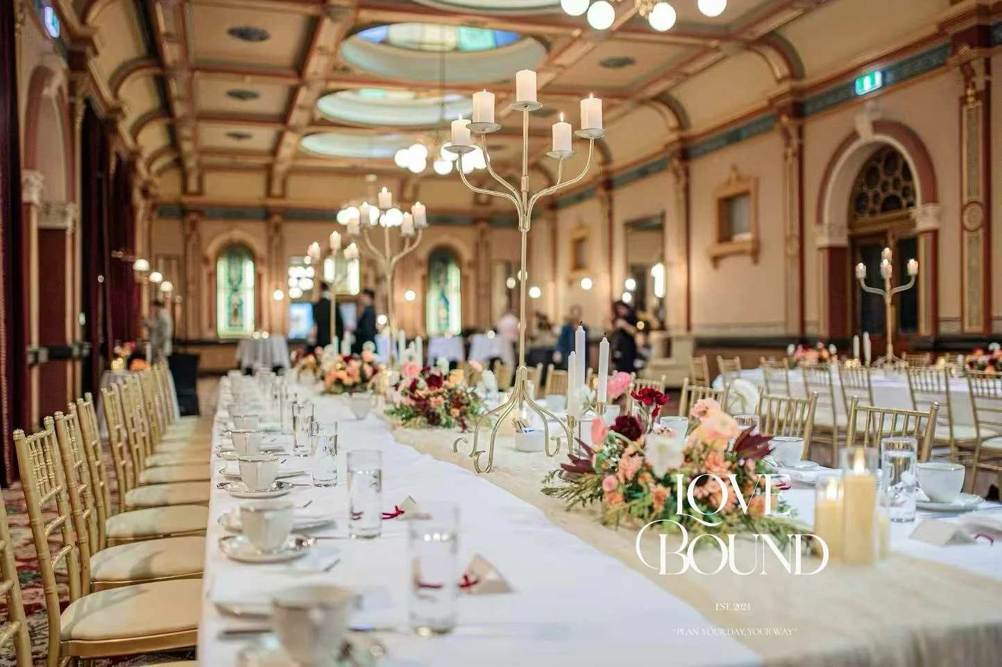 Elegant banquet hall decorated for a wedding reception with floral centerpieces, candles, and gold chairs, featuring chandeliers and stained glass windows.