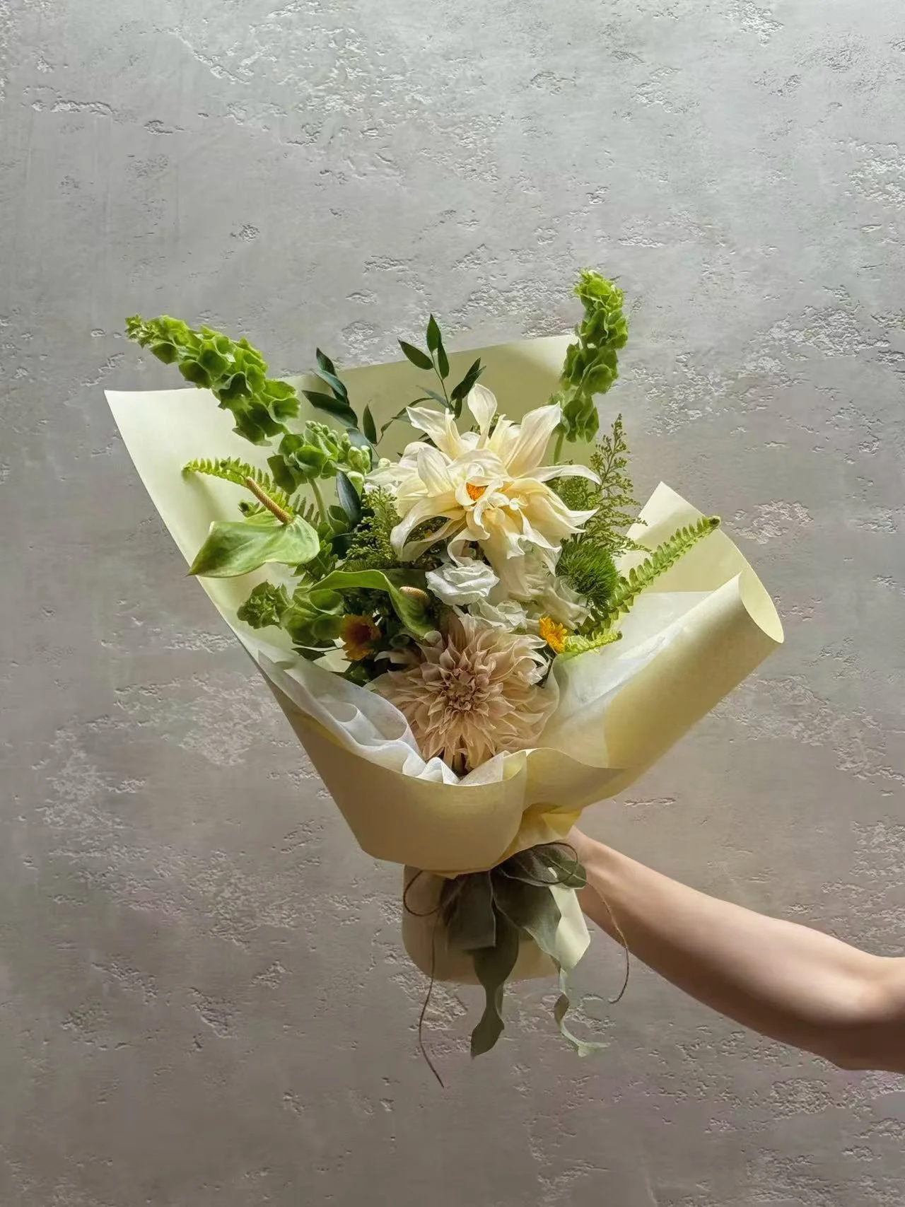 A hand holding a bouquet of white, cream, and green flowers wrapped in light-colored paper and tied with a green ribbon.