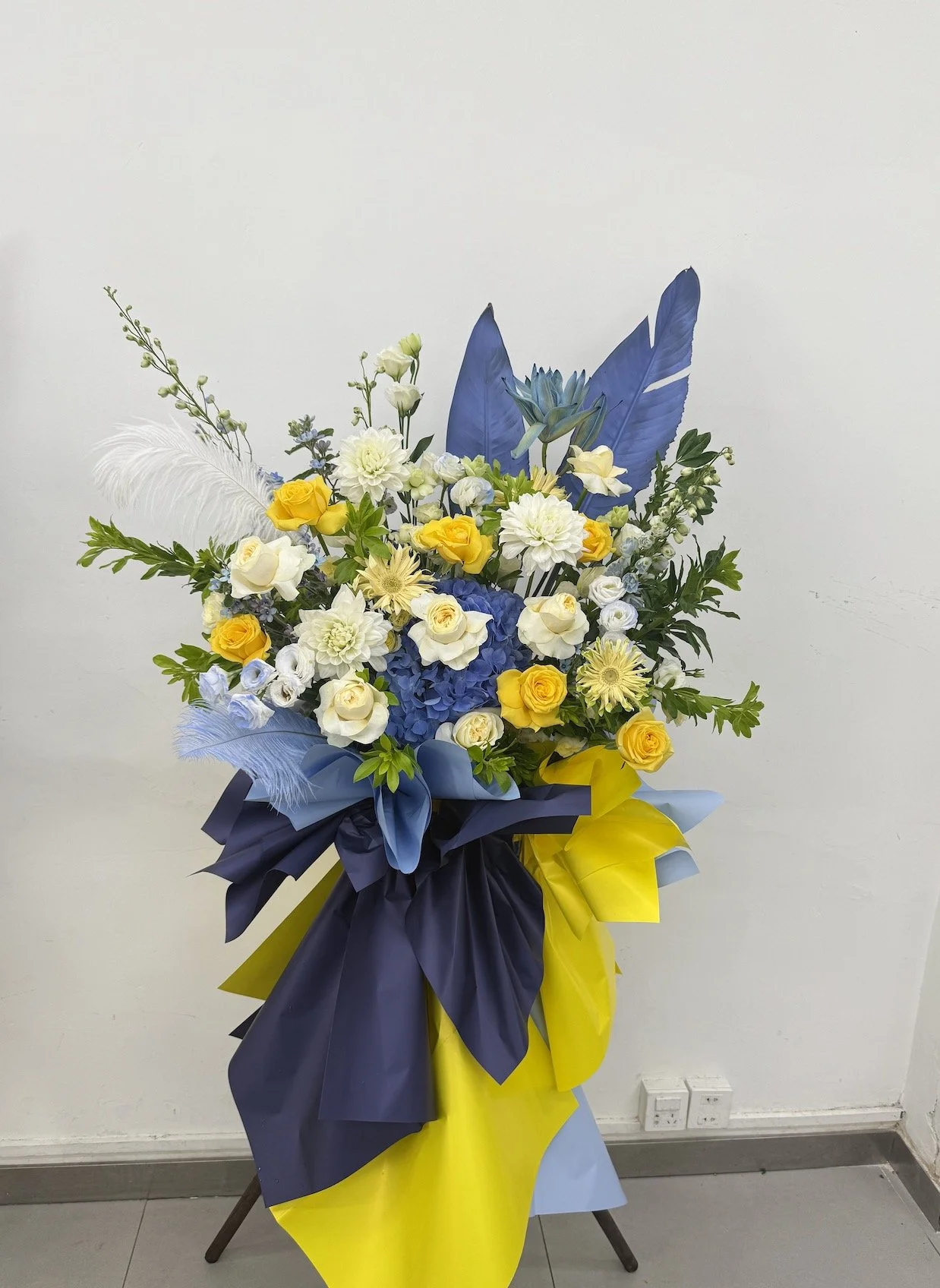 A large bouquet of flowers with white, yellow, and blue blooms, including roses and hydrangeas, decorated with green leaves and blue feathers, wrapped in blue and yellow paper.