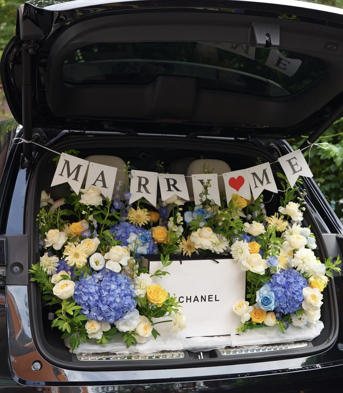 Open car trunk filled with an arrangement of white, yellow, and blue flowers, and a Chanel shopping bag, with a string of banners reading 'MARRIAGE' with a red heart in place of the 'A', hanging across the top.