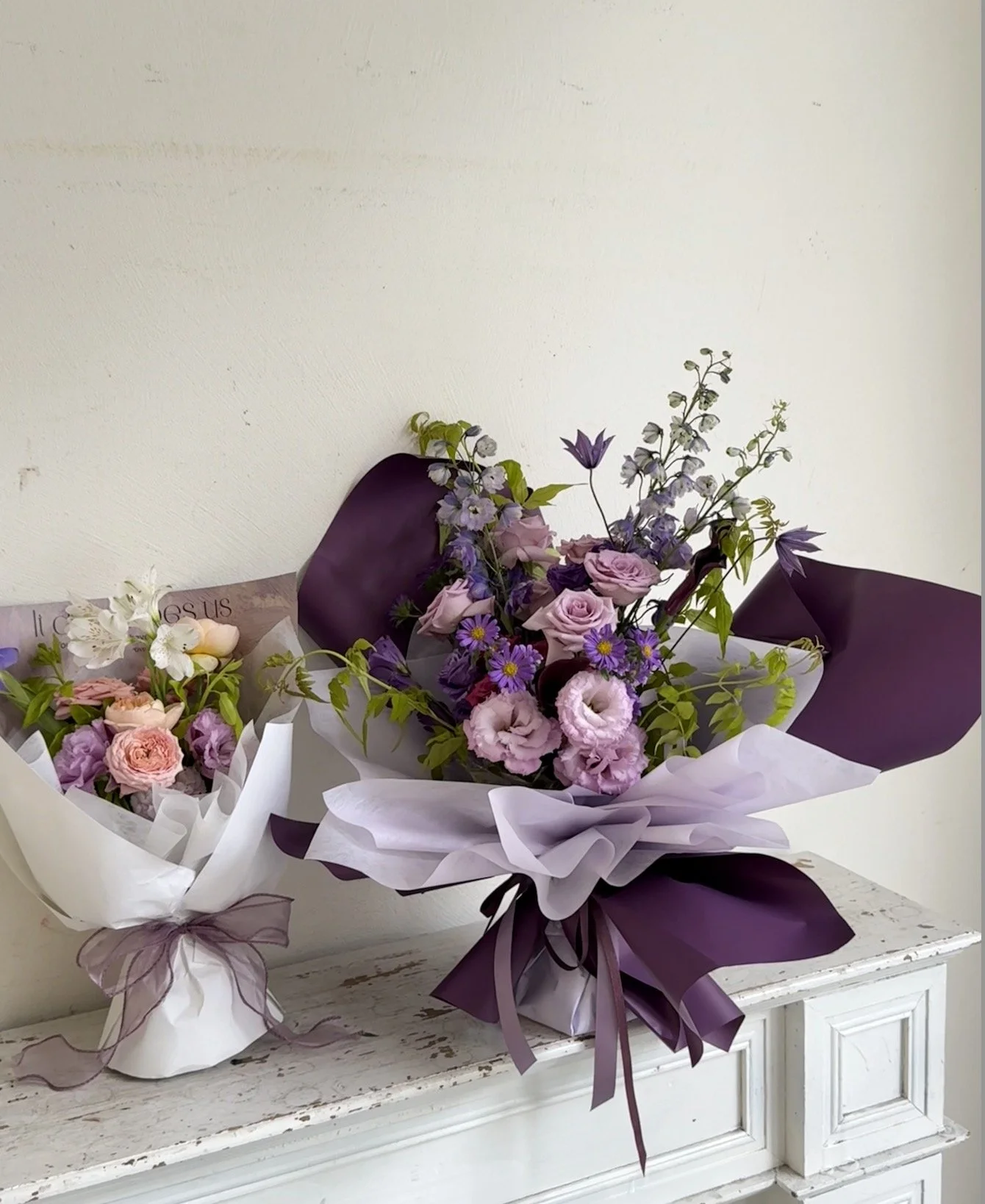 Bouquet of pink, purple, and lavender flowers wrapped in purple and white paper, tied with ribbons, placed on a white distressed dresser.
