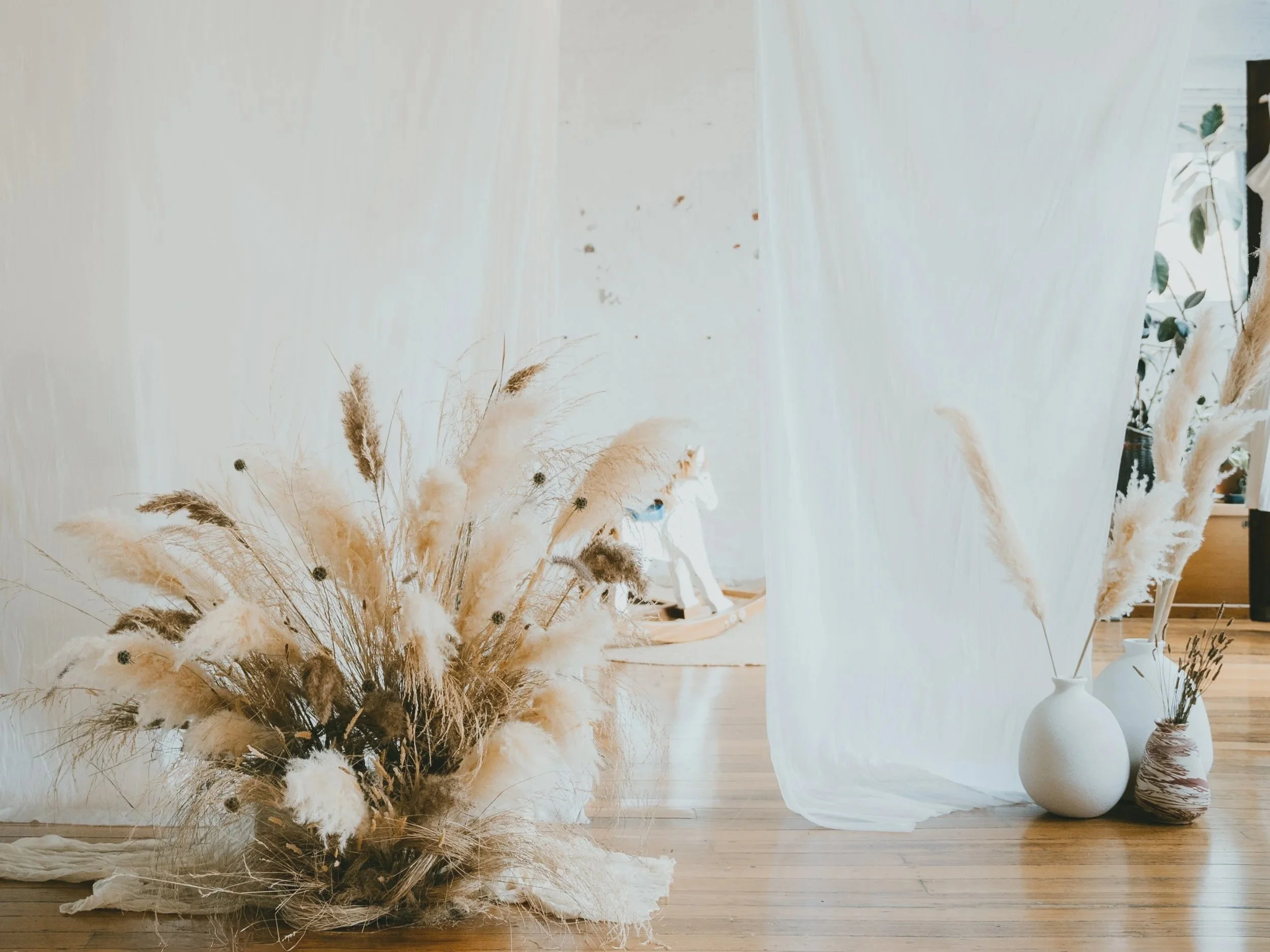 Decorative arrangement of dried pampas grass and other dried plants in white vases on a wooden floor backdrop with a sheer white curtain and natural light.