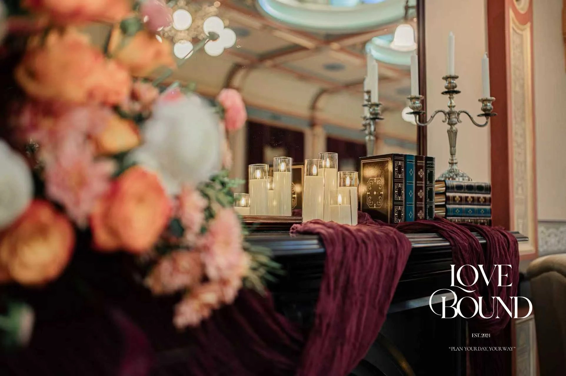 A decorated table with candle arrangements, books, and a candelabra, set against a mirror and ornate room interior, with a floral centerpiece on the left.