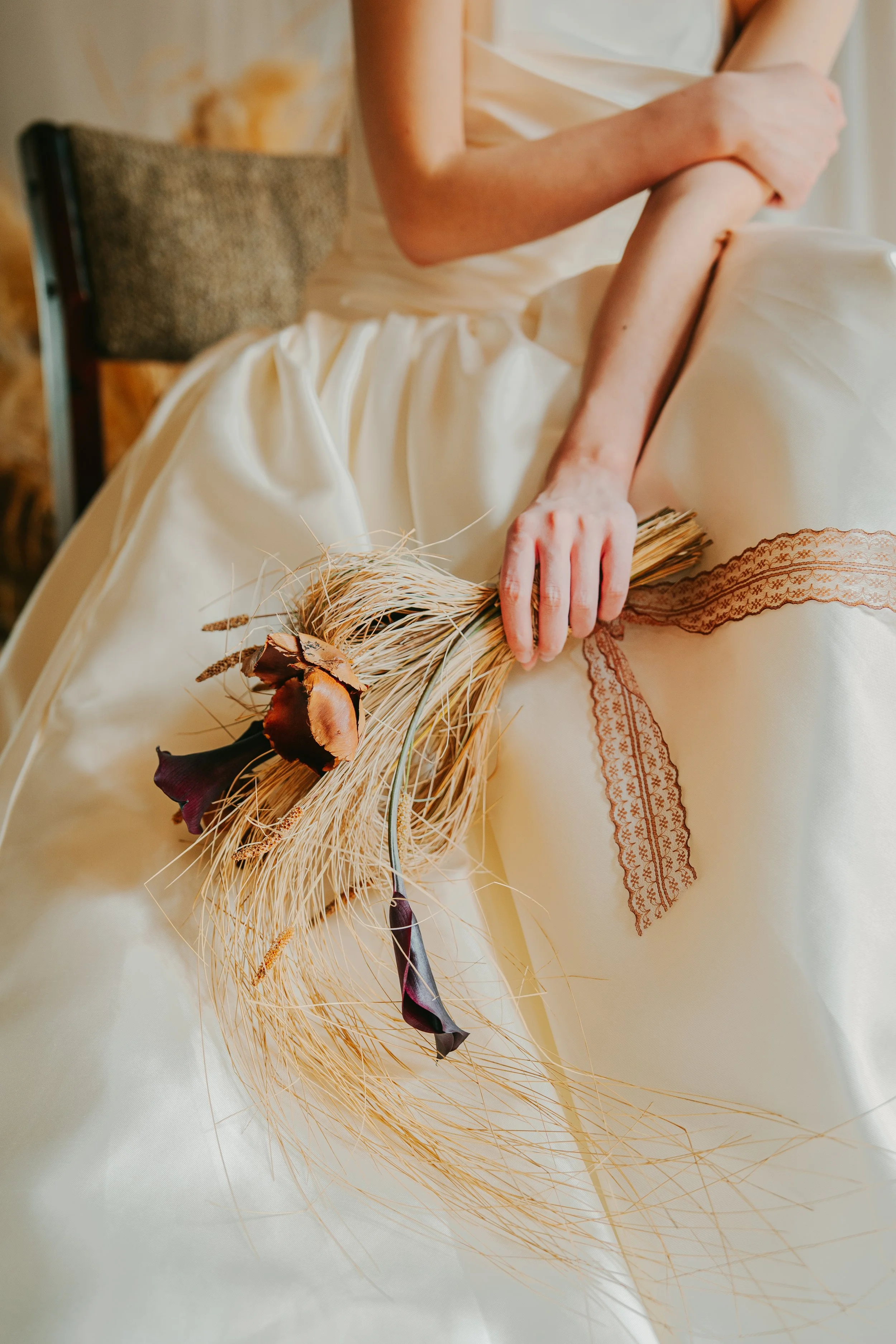 A woman in a cream-colored dress sitting on a chair, holding a dried flower bouquet with dark purple and rust-colored flowers and wheat stalks.