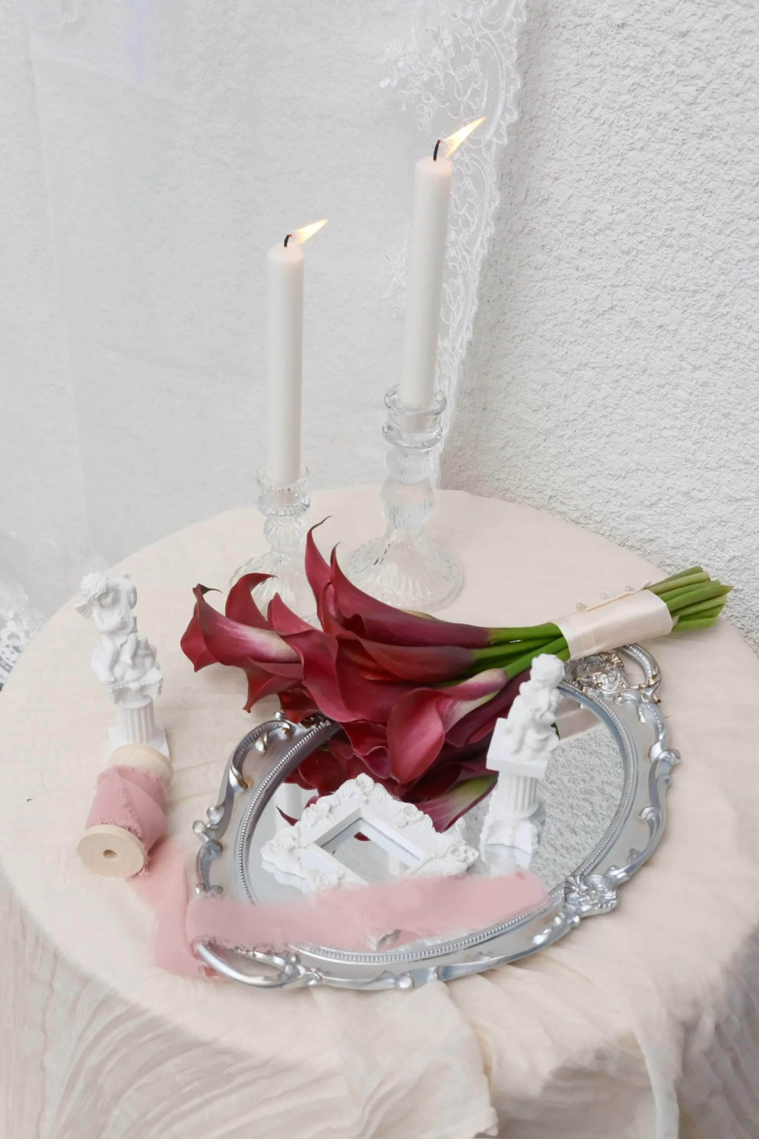 A table with two lit white candles in glass candle holders, a bouquet of red calla lilies wrapped in white paper, a vintage mirror, small white decorative sculptures of cherubs, a pink spool of thread, and a pink ruffled ribbon, with a white textured wall in the background.